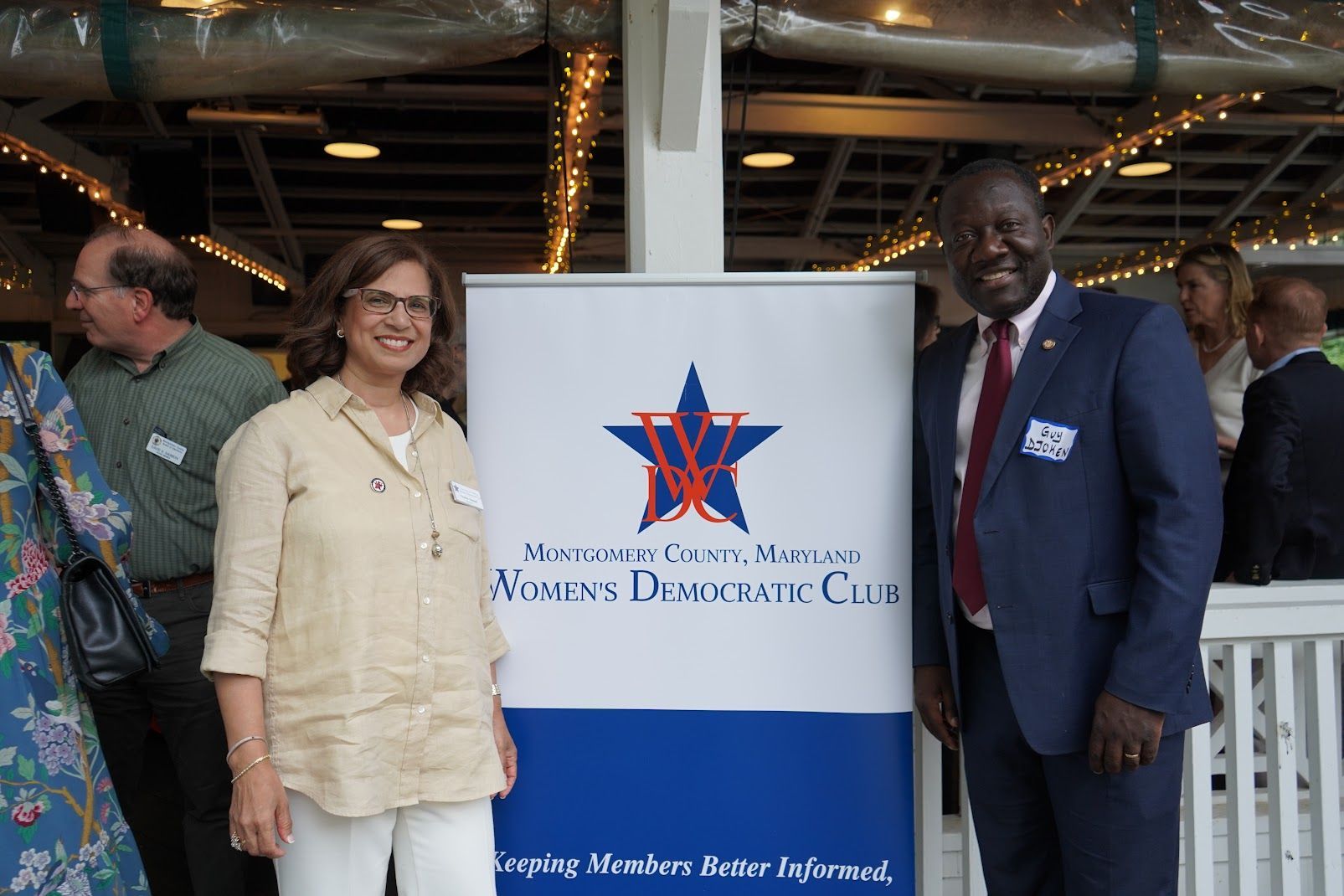 Two people stand by a sign for Montgomery County, Maryland, Women's Democratic Club. Man in suit, woman in tan shirt.