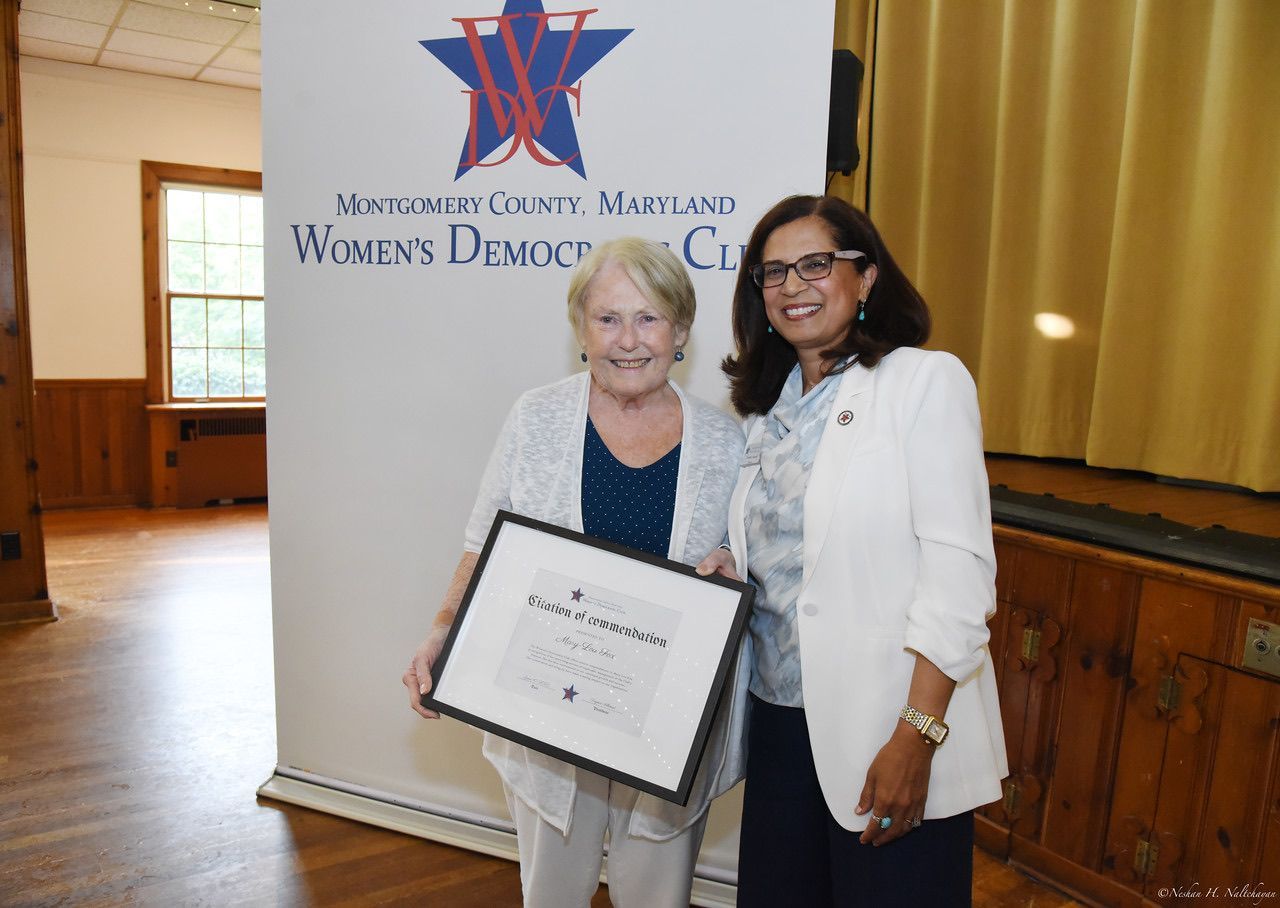 Two women smiling, one holding framed award, standing in front of a banner for a Women's Democratic Club.