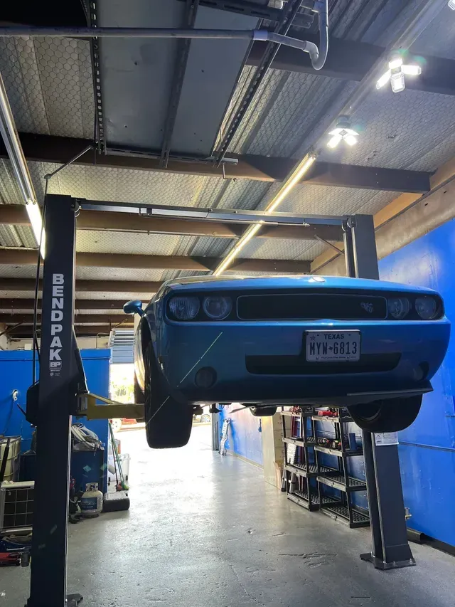 Blue classic car lifted on a shop hoist inside an auto repair garage with overhead lights  | JC Auto Repair LLC