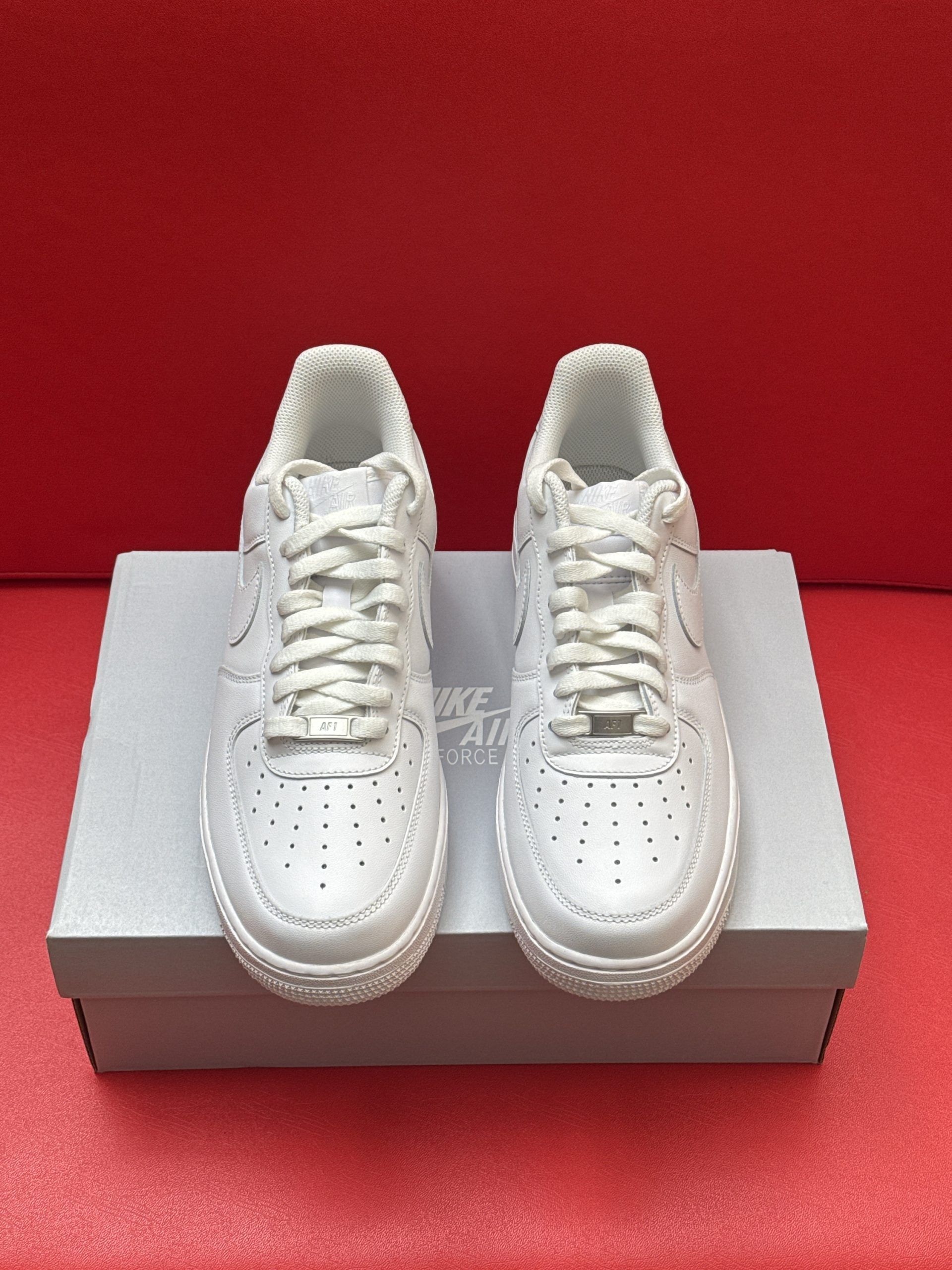 White Nike Air Force 1 sneakers, one resting on a box, the other showing sole detail; red background.