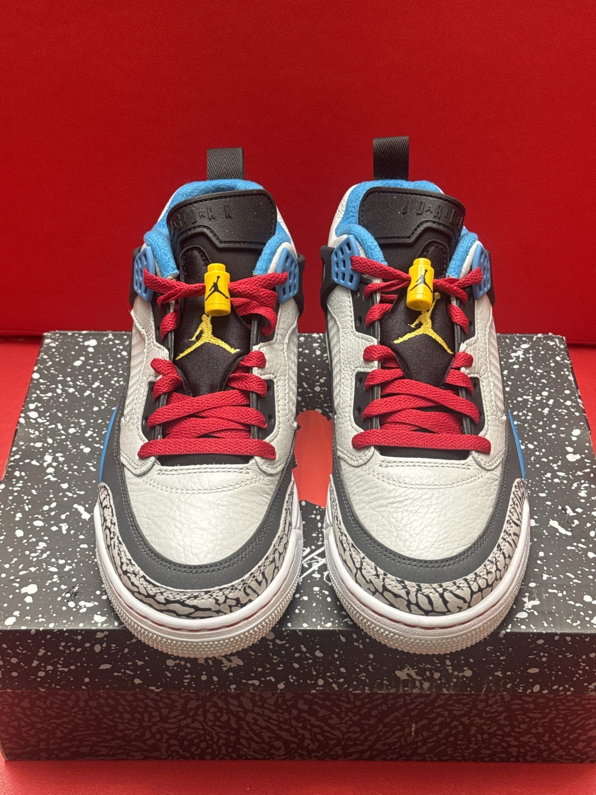 Pair of multi-colored Jordan sneakers on a shoebox against a red backdrop, showing sole detail.