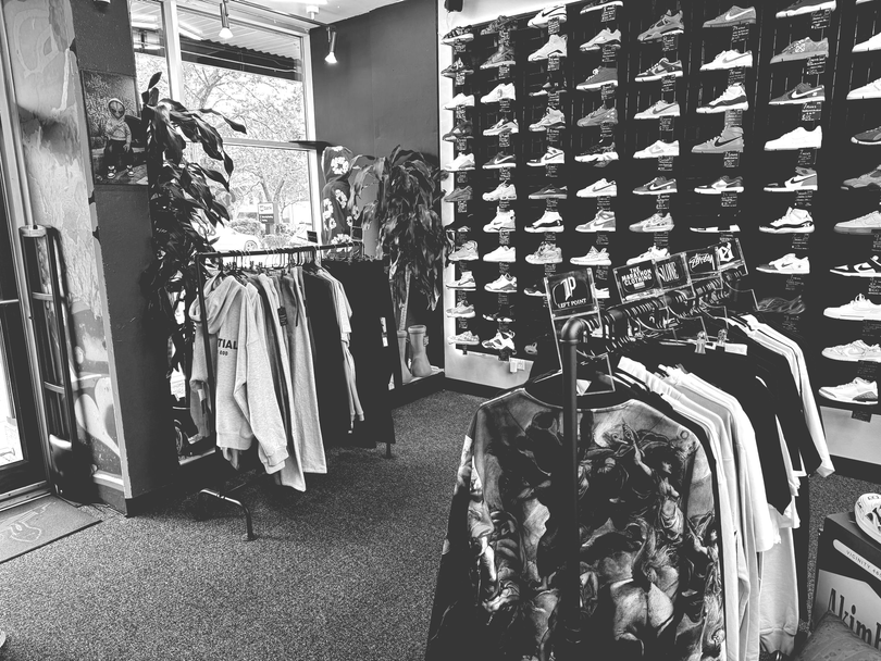Relaced sneaker store interior with shoe displays on podiums, shelves, and a black curtain. Wooden floor and palm leaves.