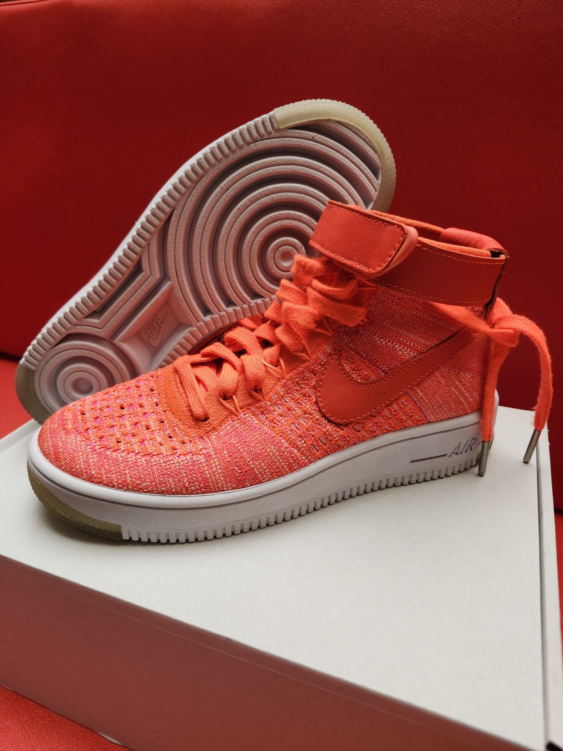 Bright orange Nike high-top sneakers on a white box, set against a red background. One shoe shows the sole.
