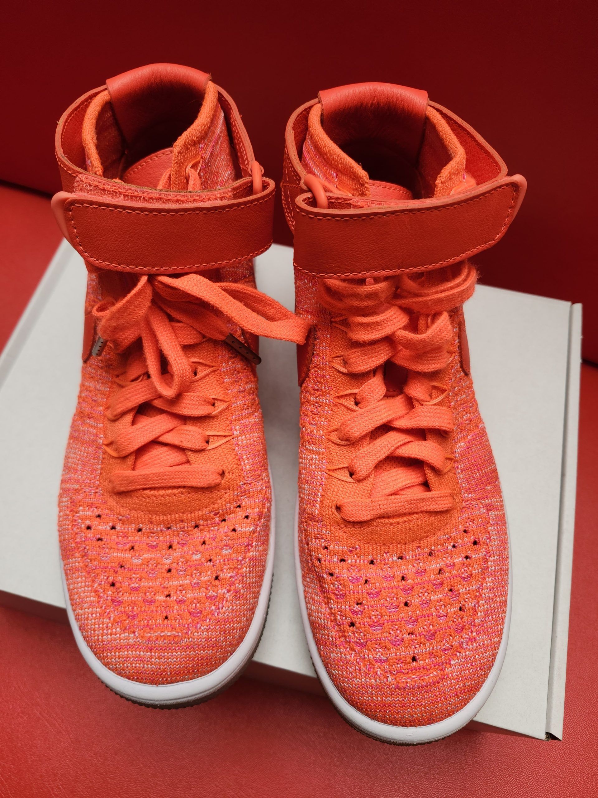 Bright orange Nike high-top sneakers on a white box, set against a red background. One shoe shows the sole.