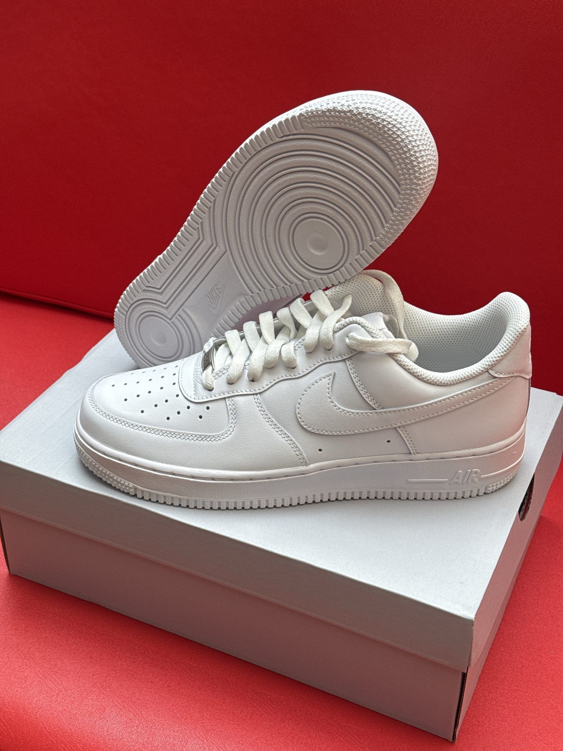 White Nike Air Force 1 sneakers, one resting on a box, the other showing sole detail; red background.