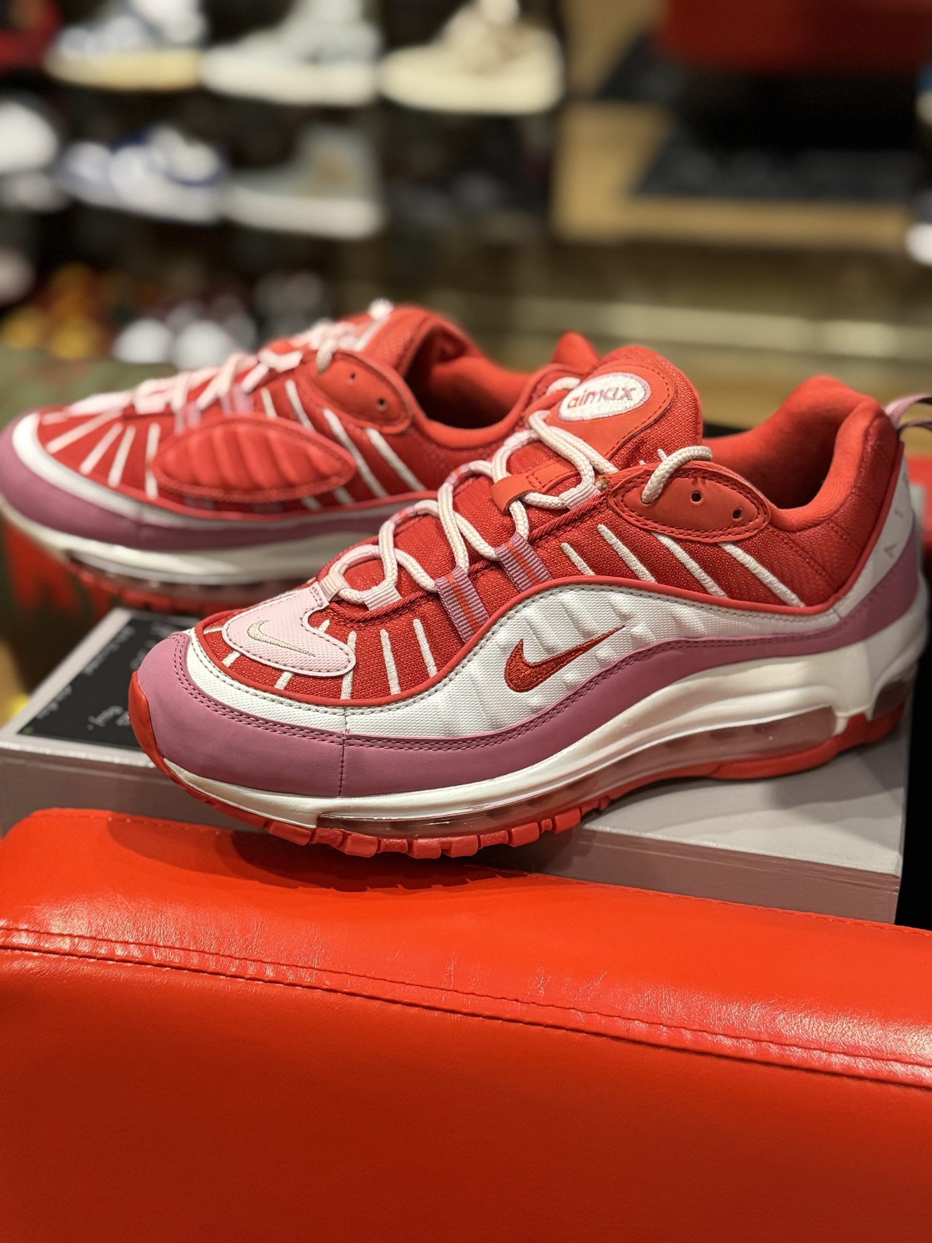 Red Nike Air Max 98 sneakers on a red surface, with a white and pink sole and laces.