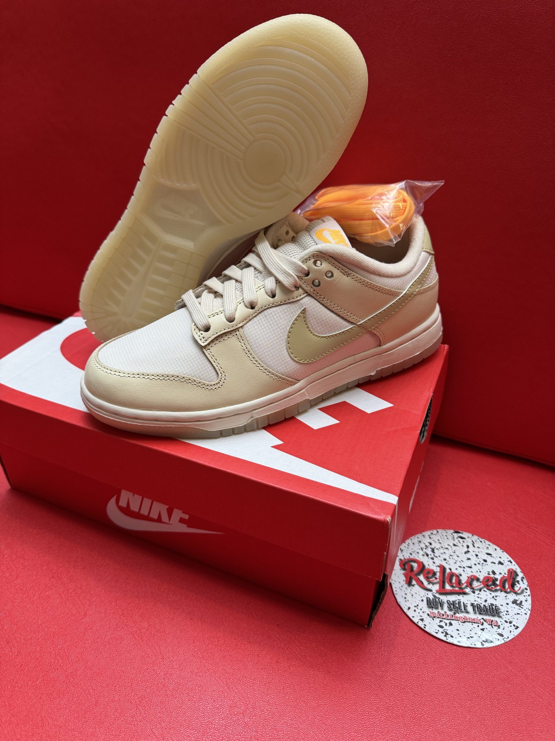 Beige Nike Dunk Low sneaker with orange laces, box, and sole on a red surface.