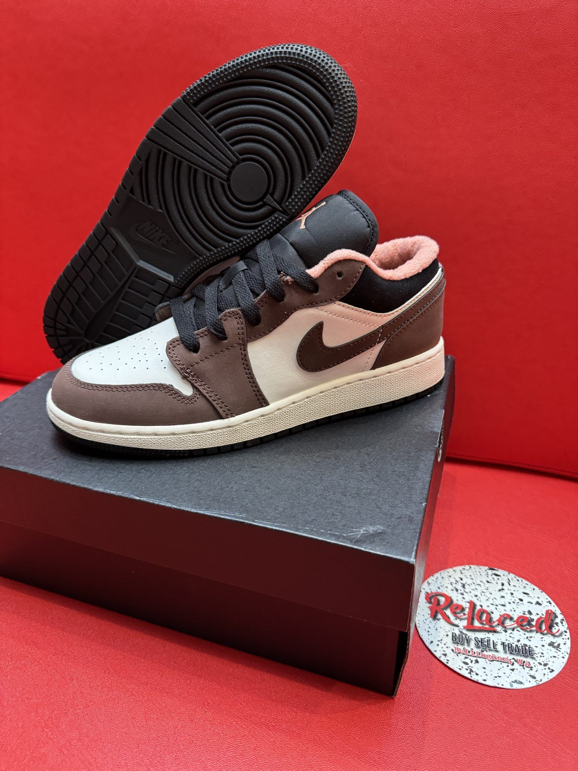 A small, brown, and cream-colored sneaker rests on a black box, with a sole in view on a red surface.