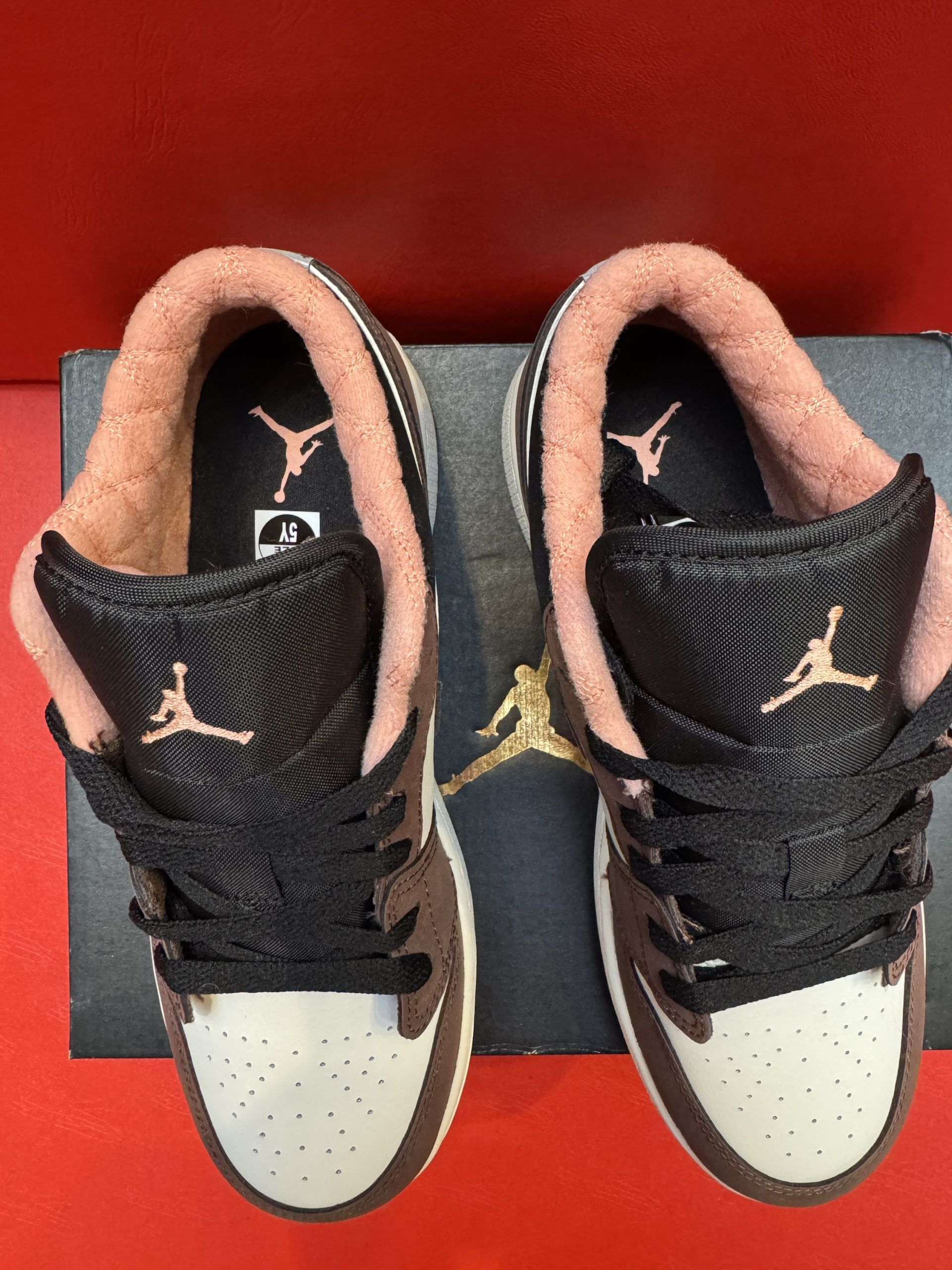 A small, brown, and cream-colored sneaker rests on a black box, with a sole in view on a red surface.
