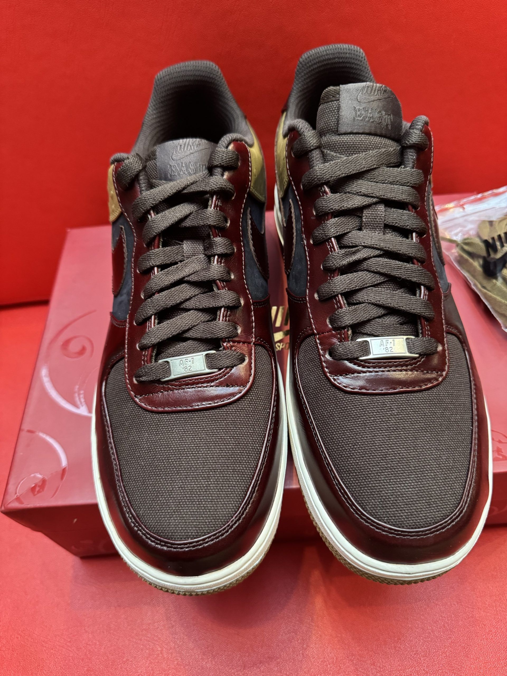 Air Force 1 sneakers on a red box, featuring brown, navy, and tan colors with a gum sole and extra laces.