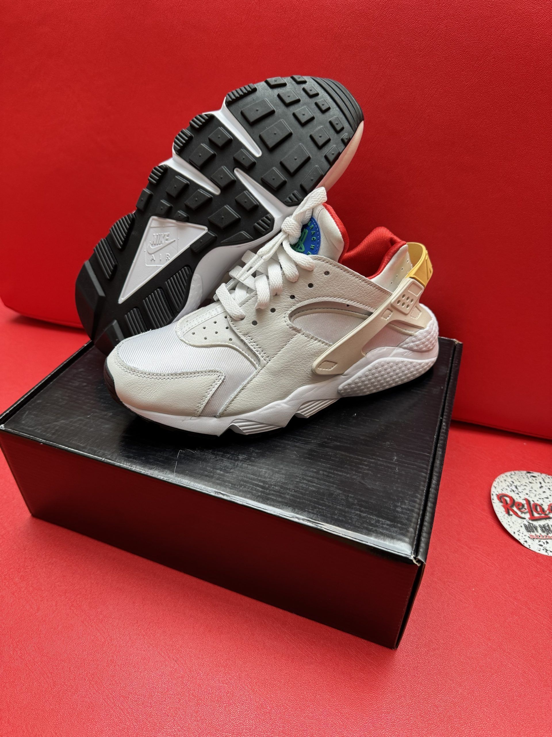 White and tan Nike Huarache sneakers on a black box, one shoe up, red background.