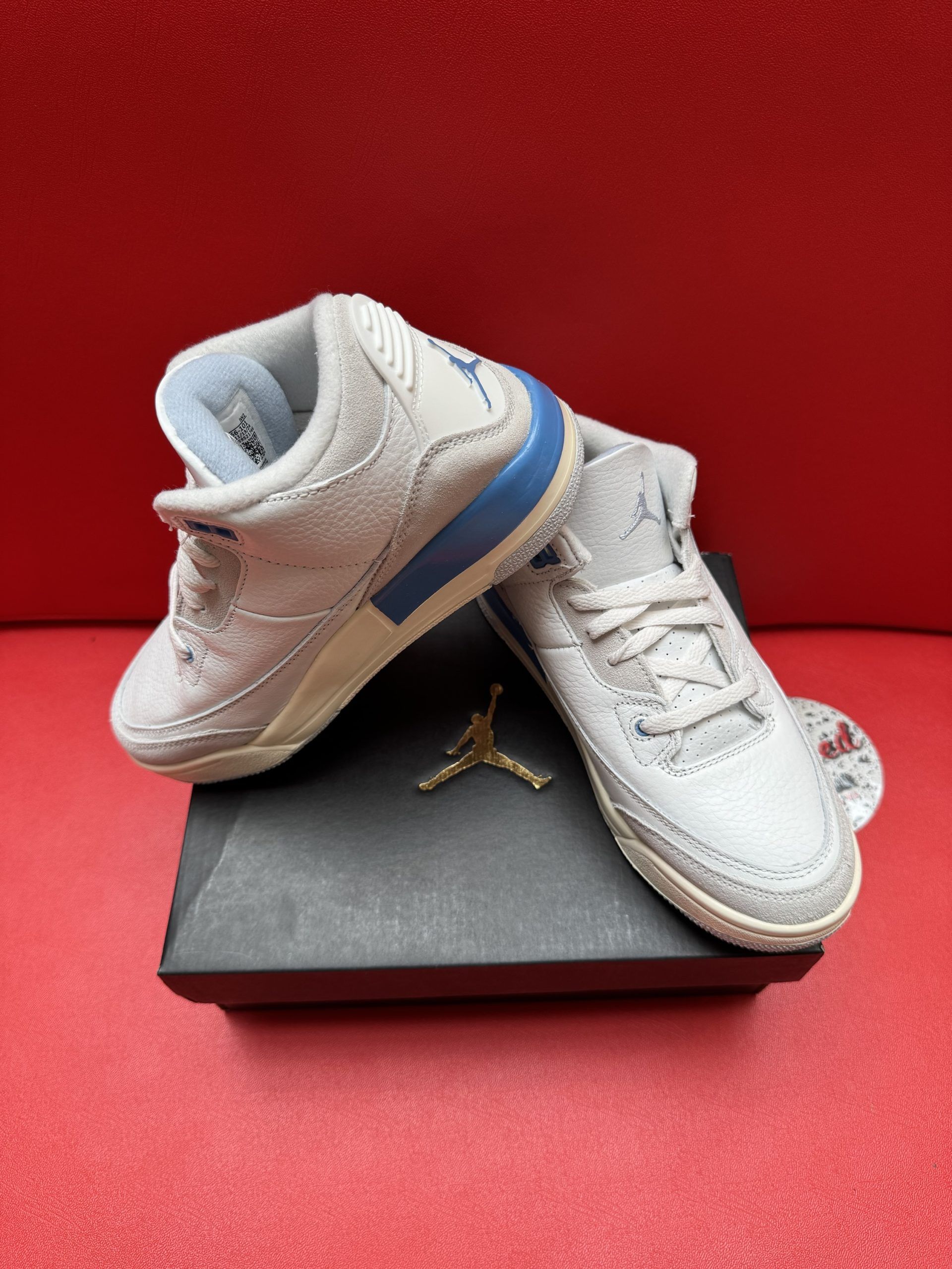 Air Jordan 3 sneakers, light grey, baby size, on a black box, with a red background.