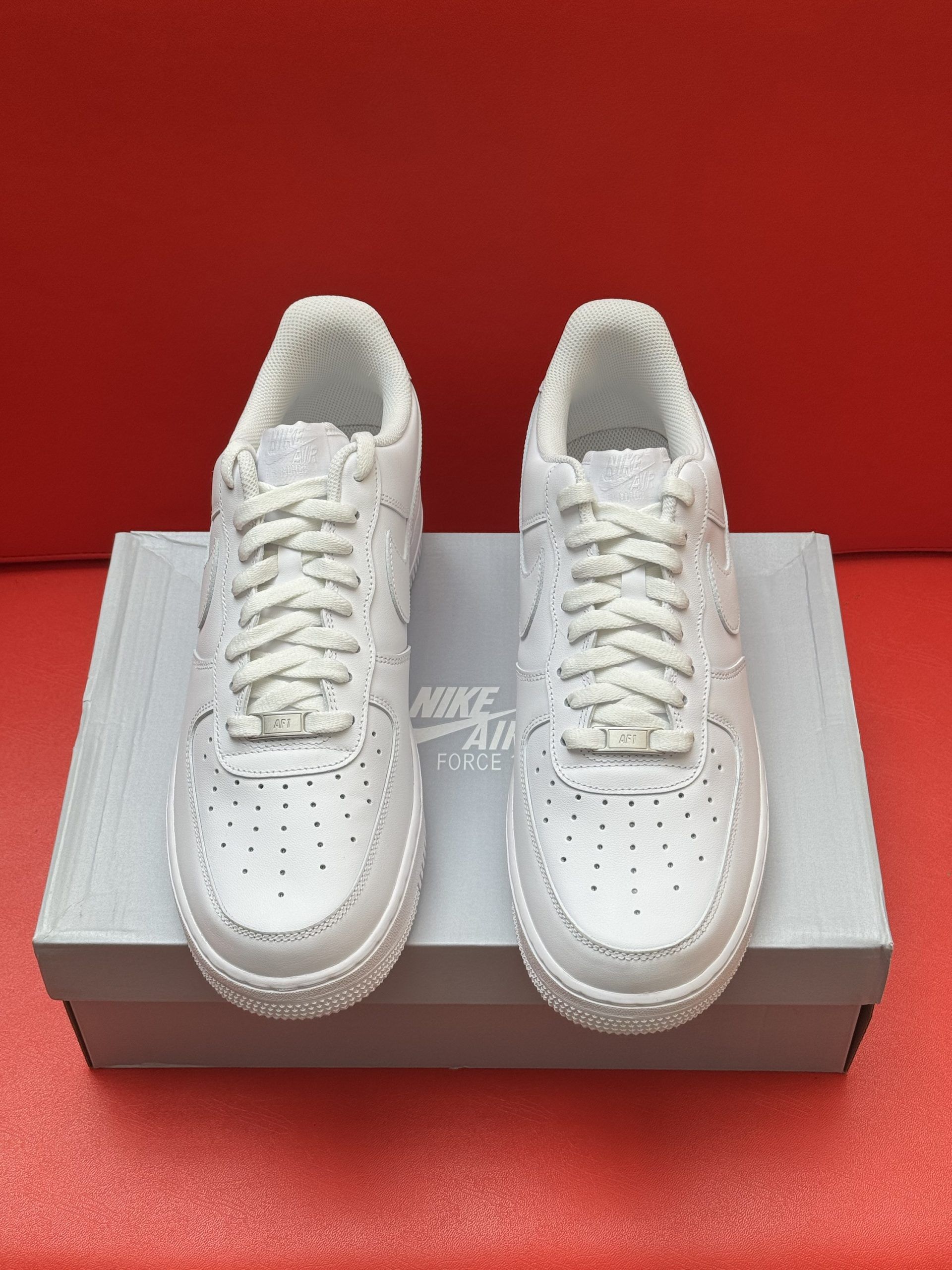 White Nike Air Force 1 sneakers on a box, sole up, against a red background.