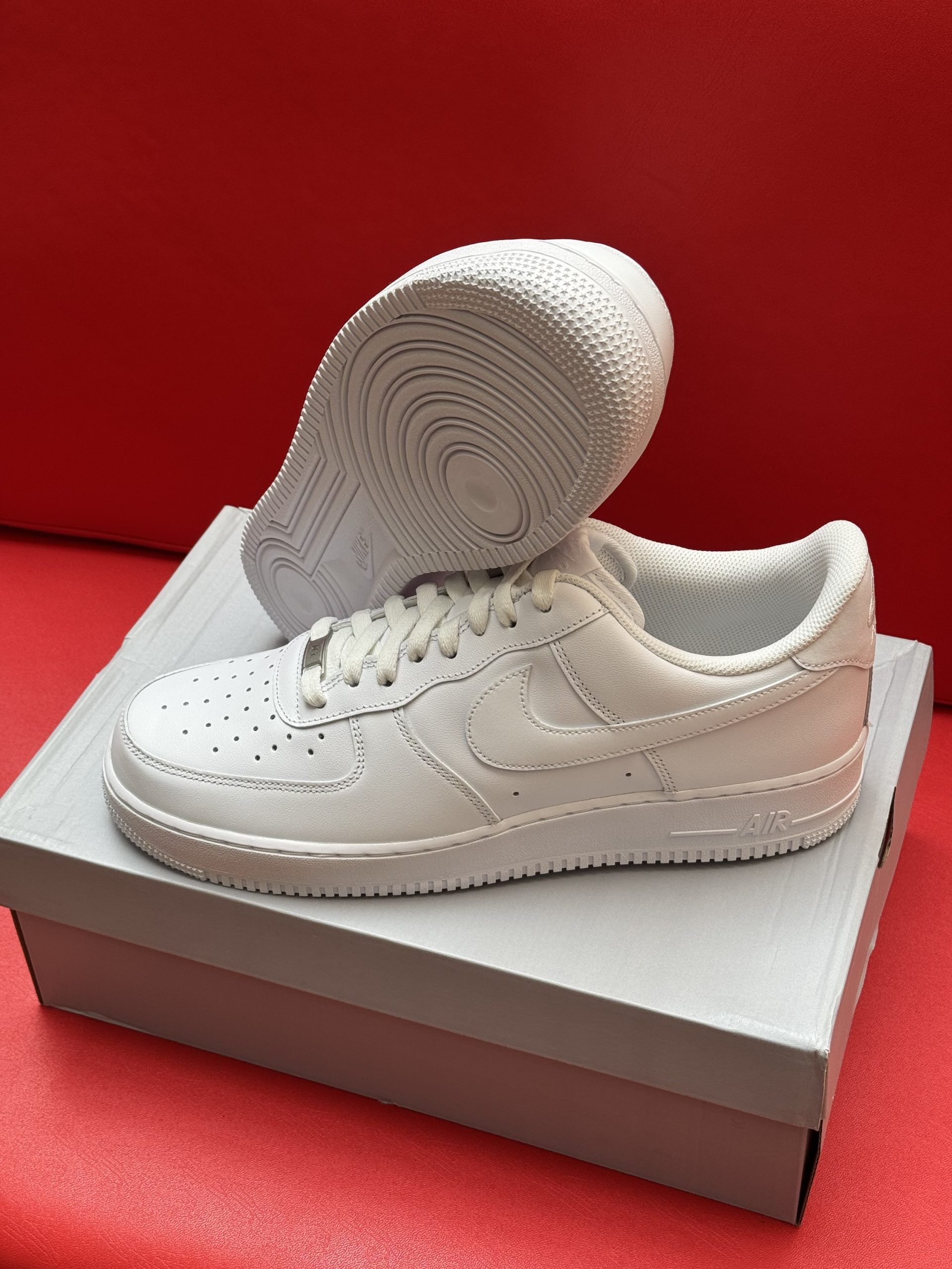 White Nike Air Force 1 sneakers on a box, sole up, against a red background.