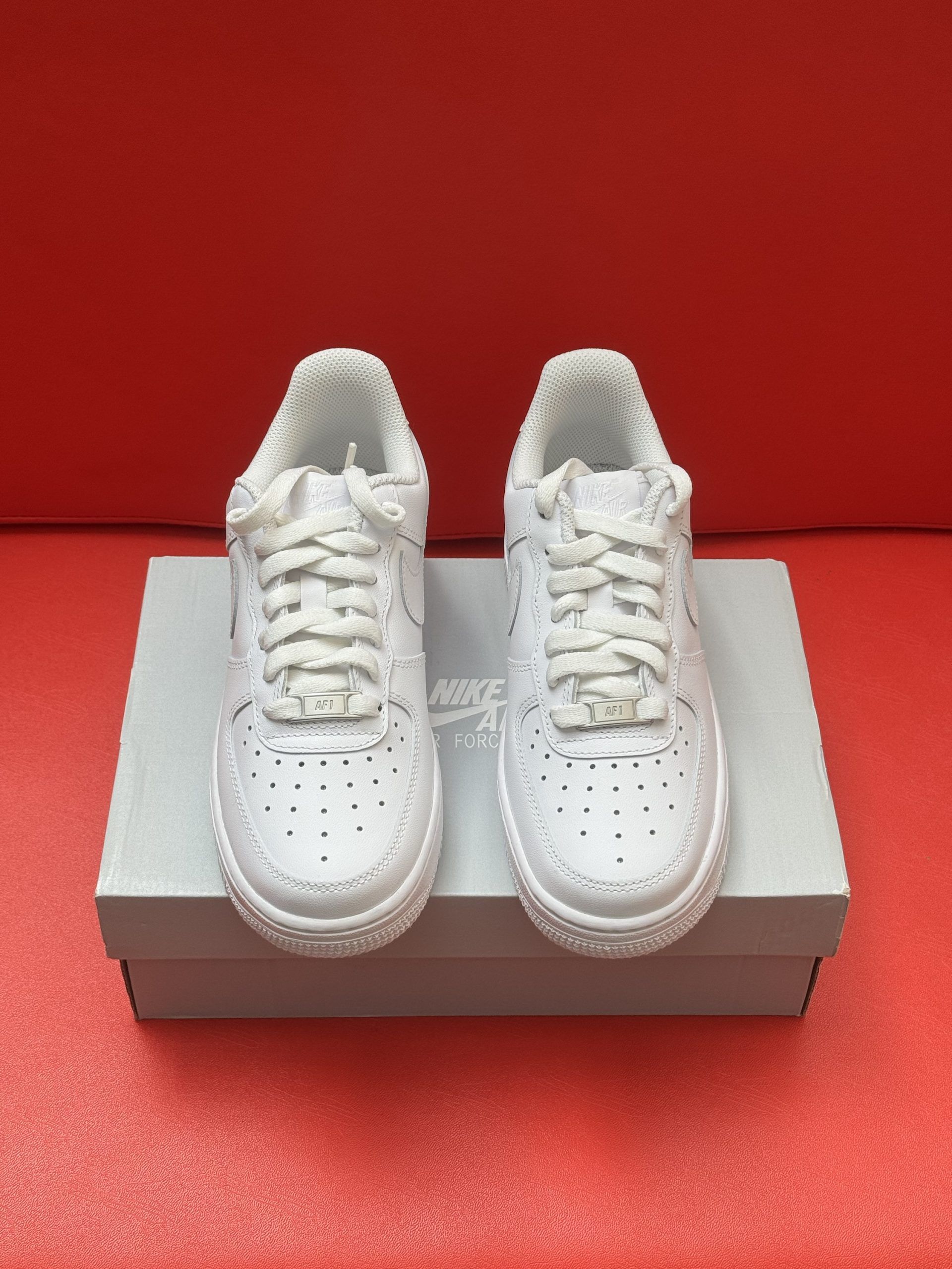 White Nike Air Force 1 sneakers on a box, one flipped to show sole, against a red background.