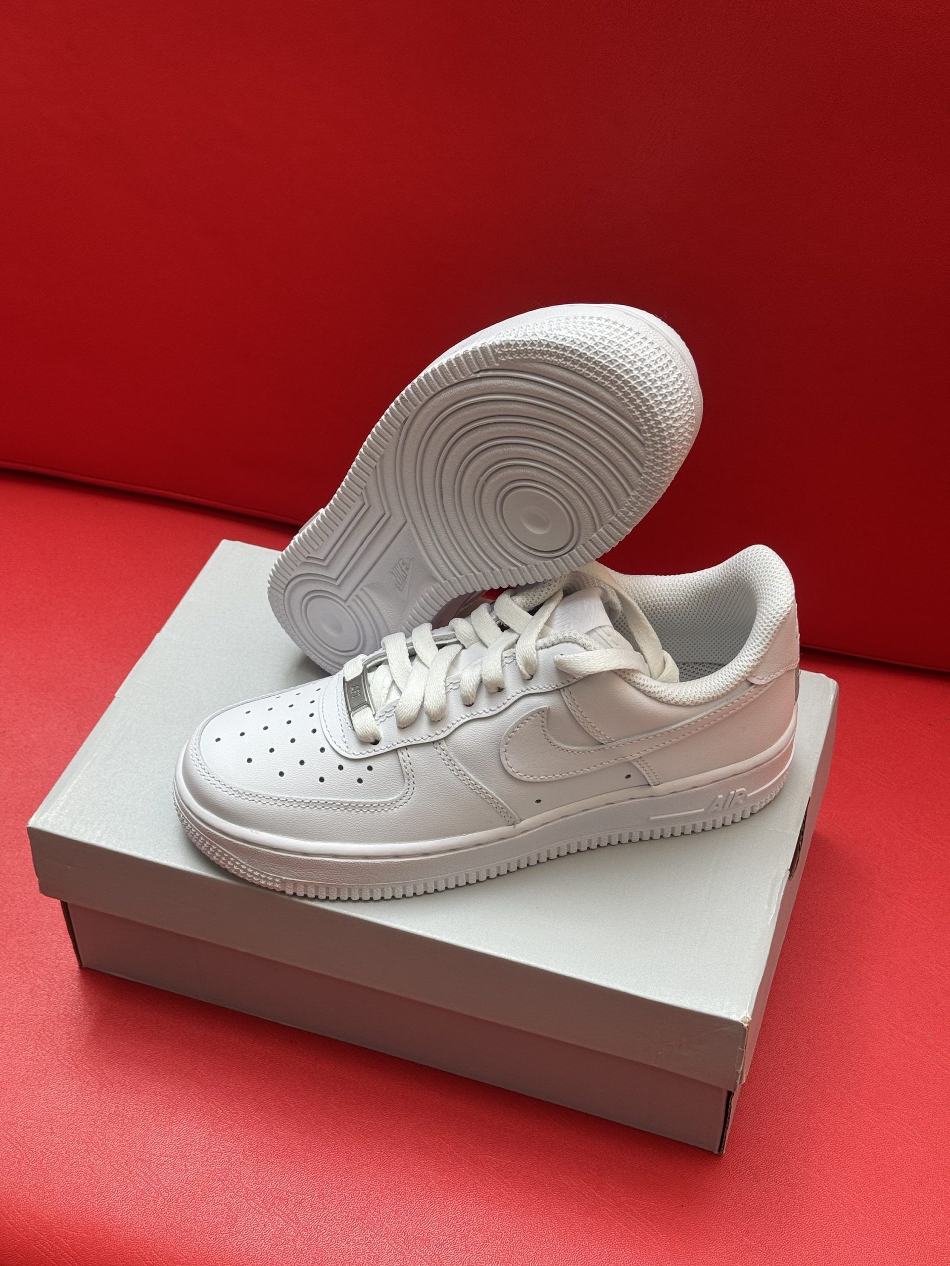 White Nike Air Force 1 sneakers on a box, one flipped to show sole, against a red background.