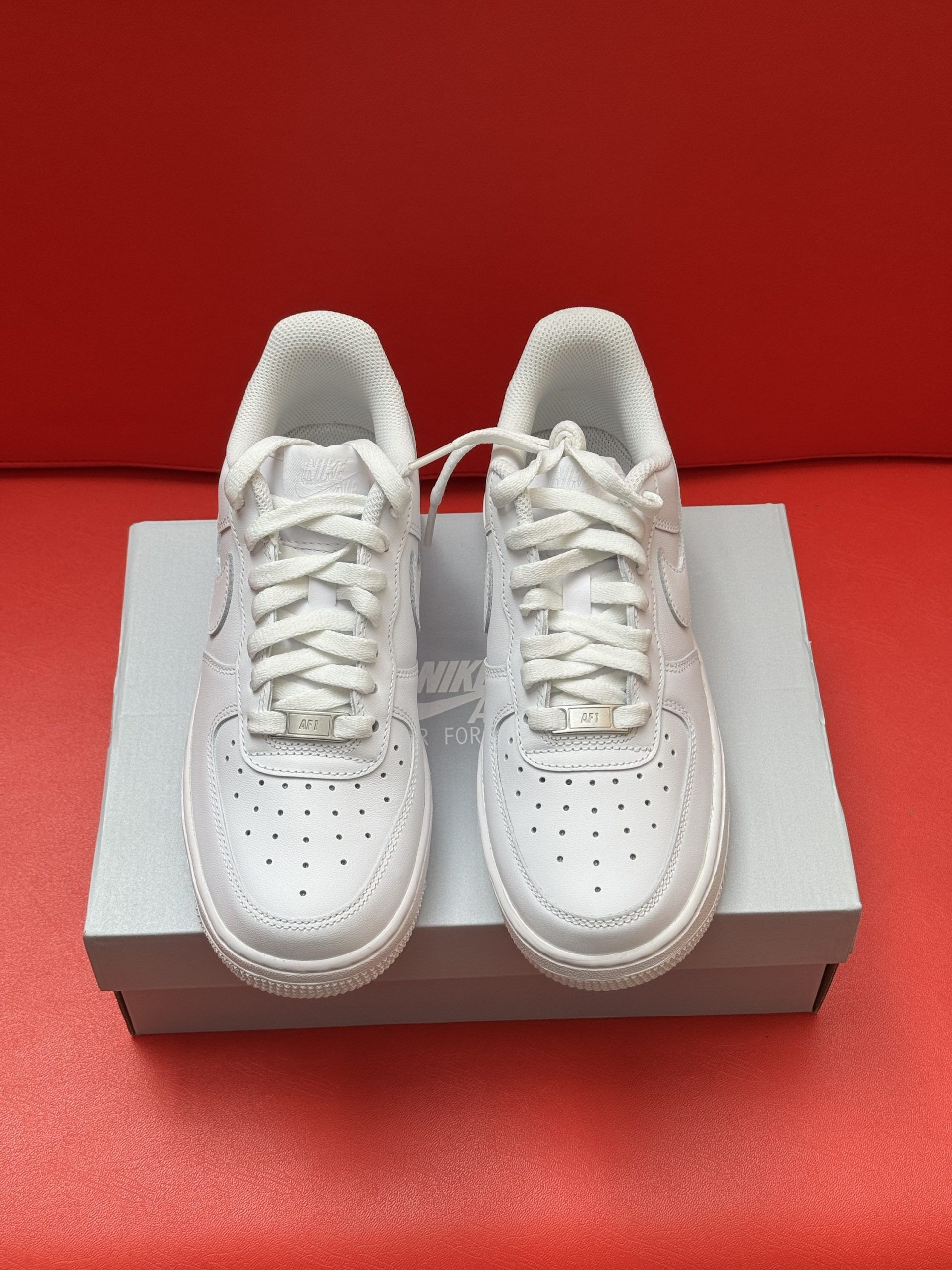 White Nike Air Force 1 sneakers on top of a shoe box, with the sole of one shoe visible. Red background.