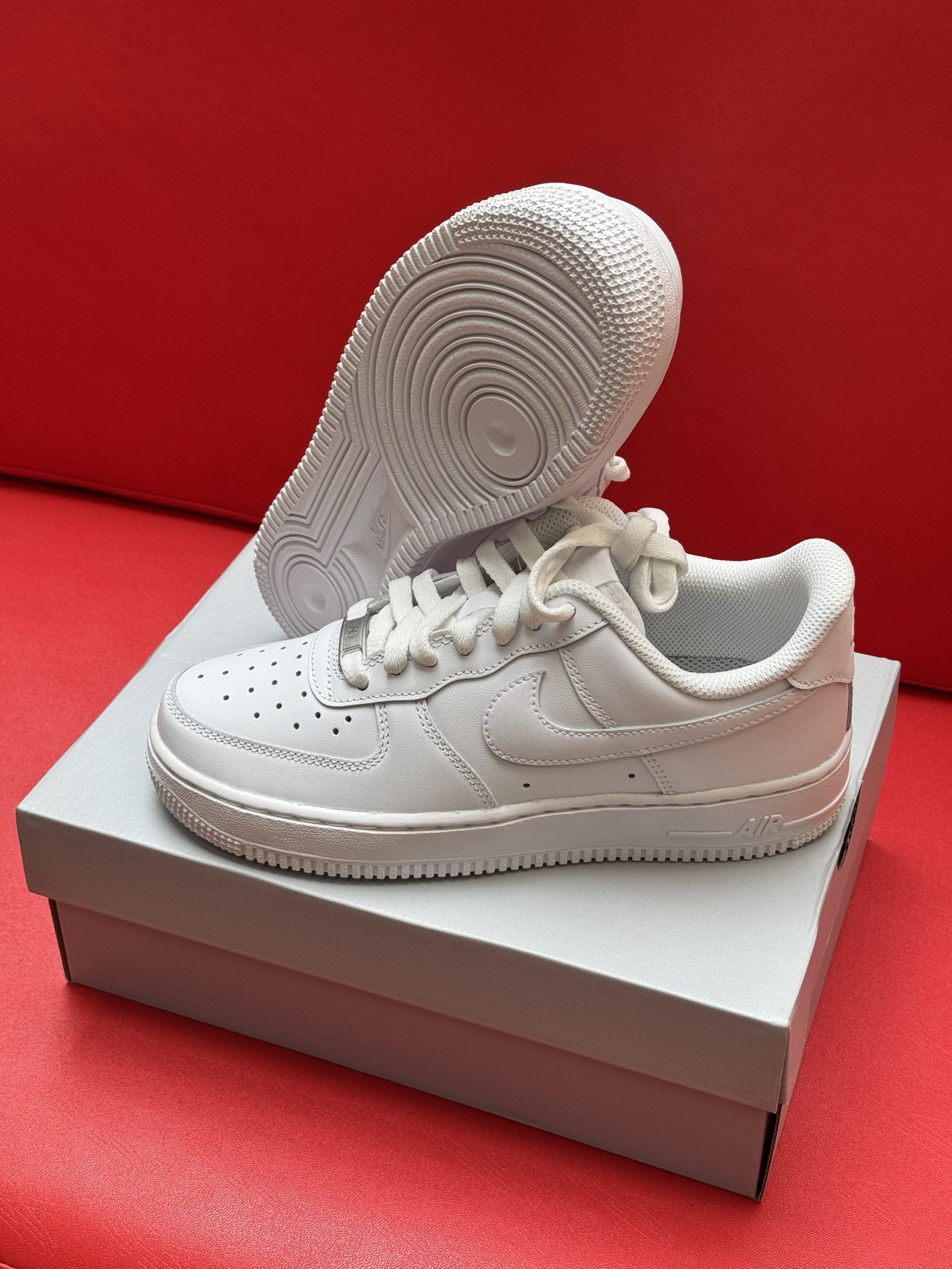 White Nike Air Force 1 sneakers on top of a shoe box, with the sole of one shoe visible. Red background.