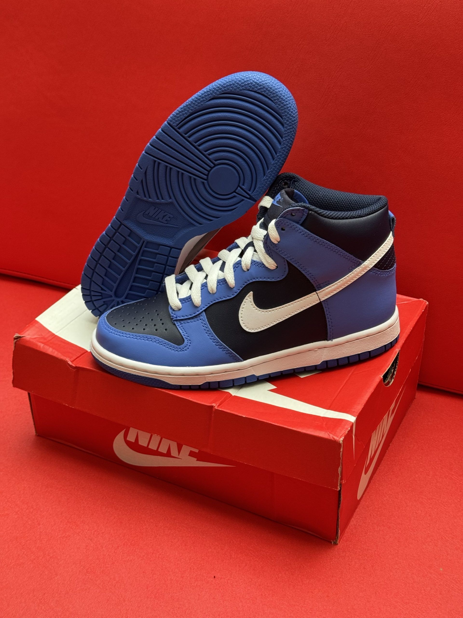 Blue and black Nike Dunk high-top sneaker on a red Nike box, with the blue sole visible, set against a red background.