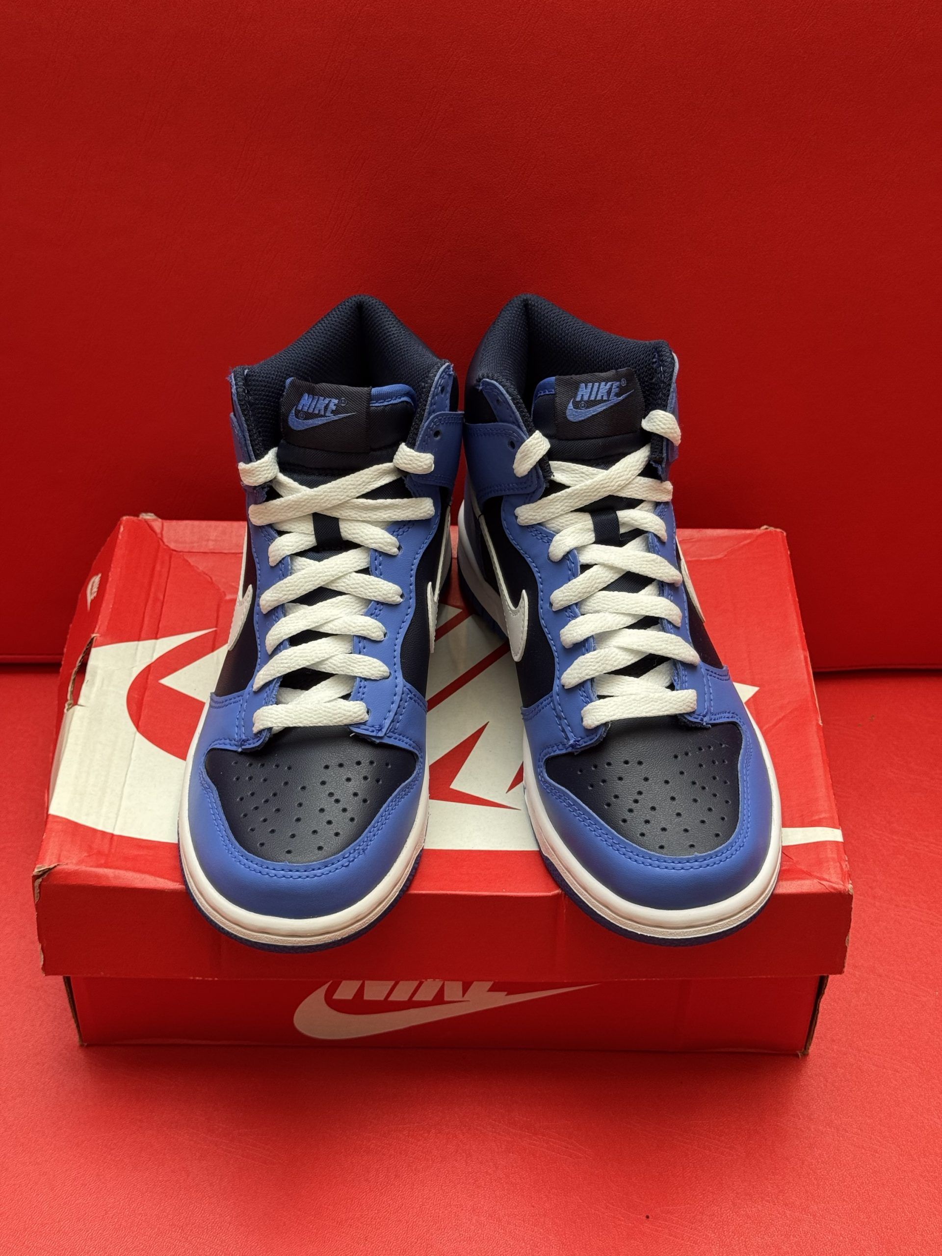 Blue and black Nike Dunk high-top sneaker on a red Nike box, with the blue sole visible, set against a red background.