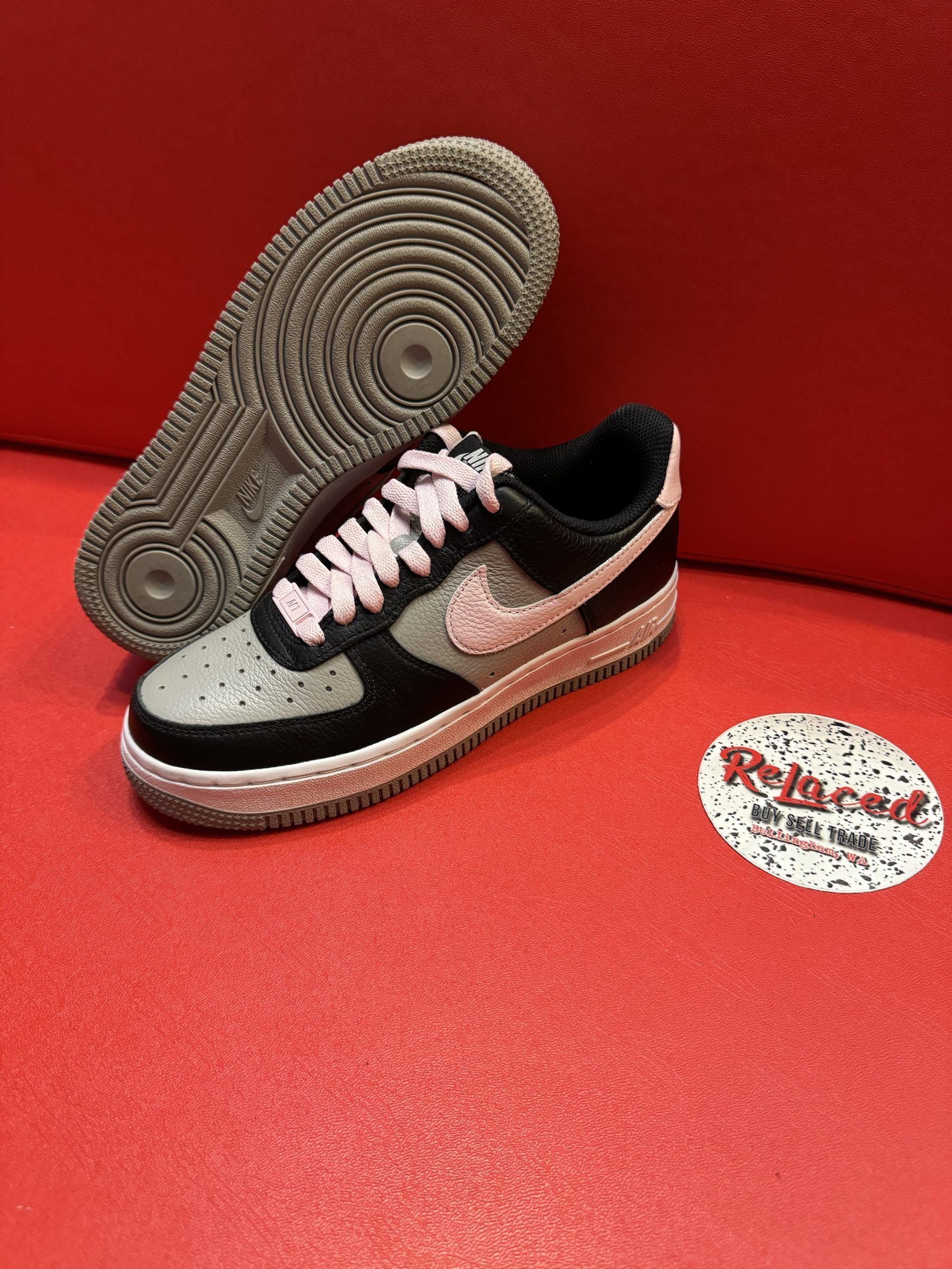 Black, gray, and pink Nike Air Force 1 sneaker with light pink laces, sole visible, on red surface.