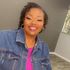 Smiling person in a denim jacket and pink top standing indoors with a gray wall and carpeted floor.