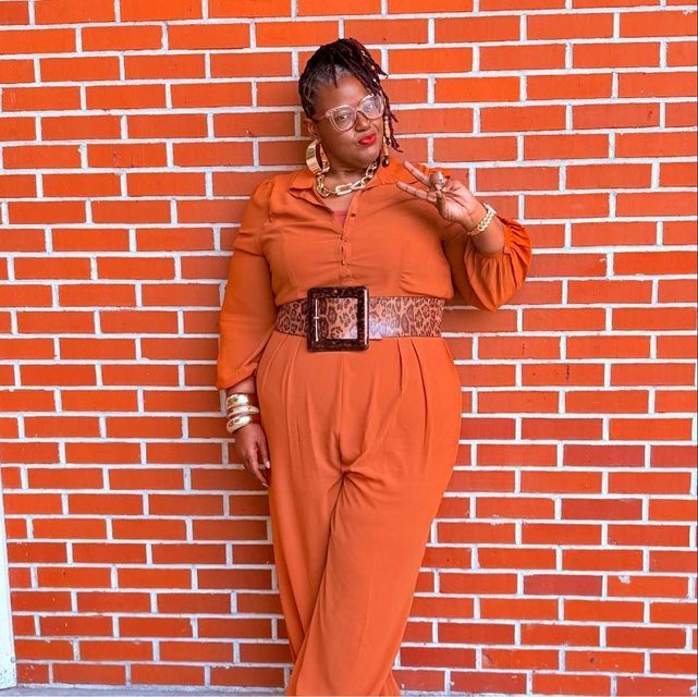 Person in orange outfit posing against a red brick wall, holding up a peace sign.