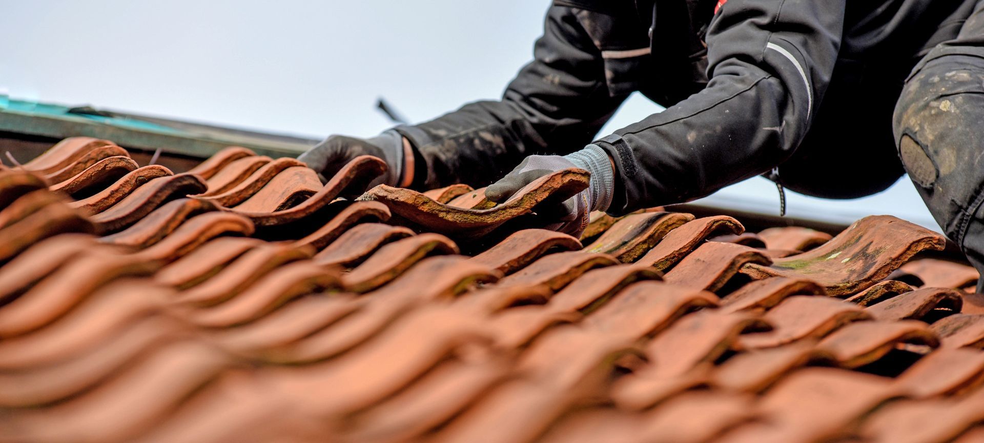 Person wearing gloves working on a red-tiled roof, with clear sky in the background.