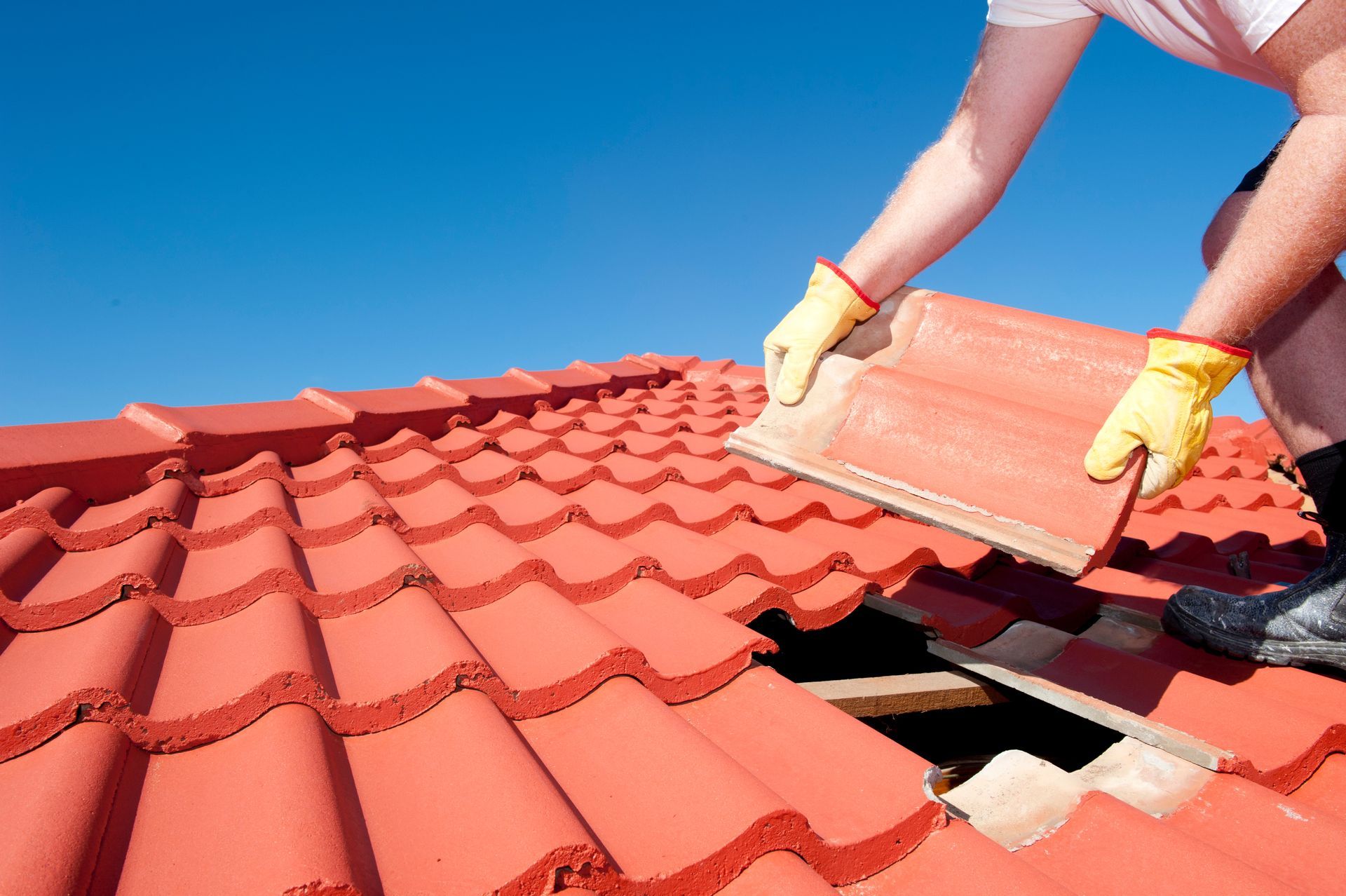 View of the hands of a roofing worker repairing a roof tile against a blue sky.