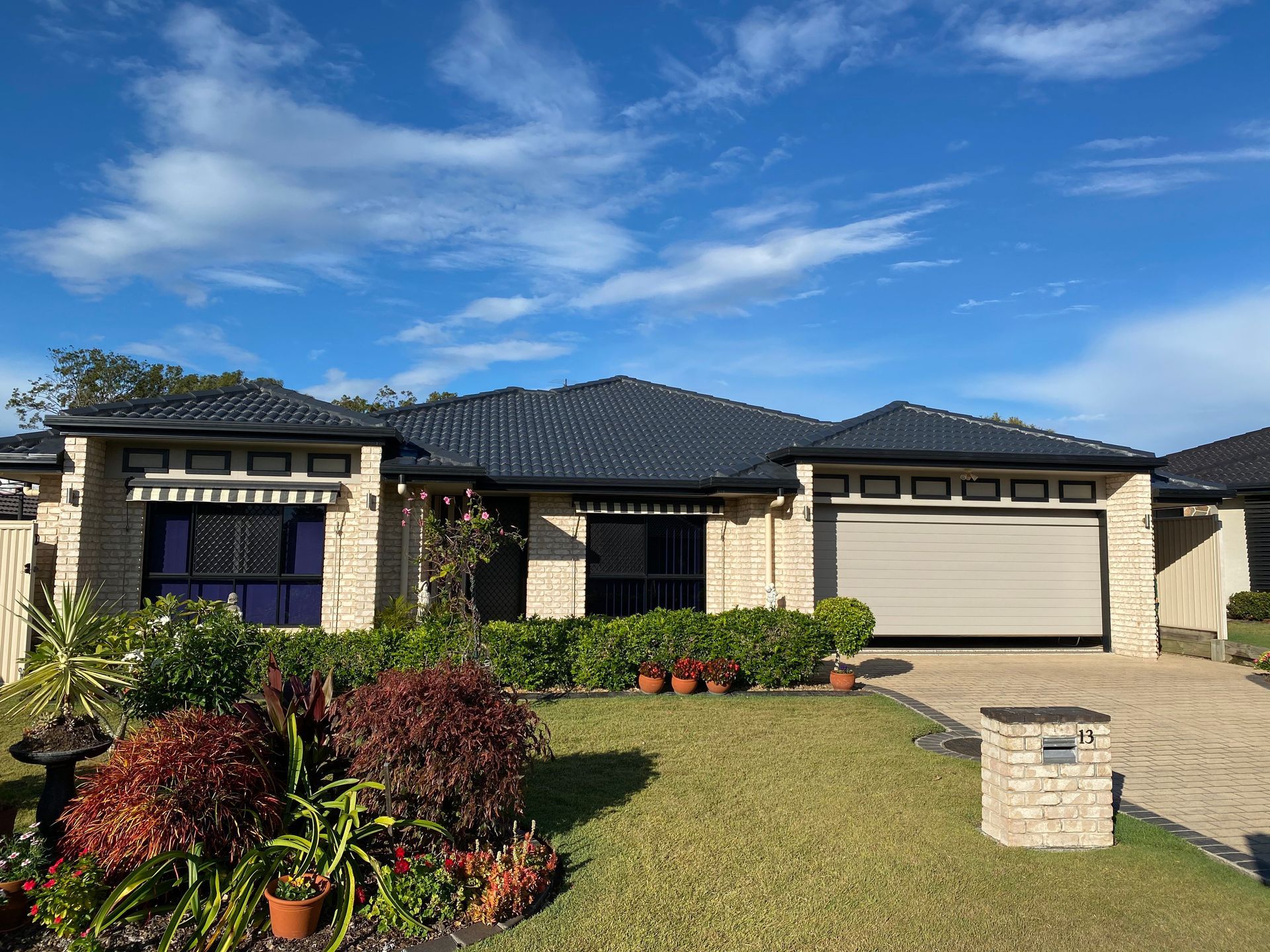 Beige brick house with blue roof and garage, lawn, plants, and blue sky.