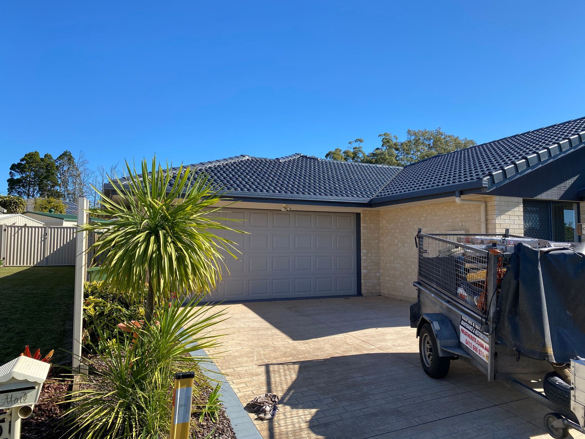 A house with a blue tiled roof, garage, and trailer parked on a paved driveway under a clear blue sky.