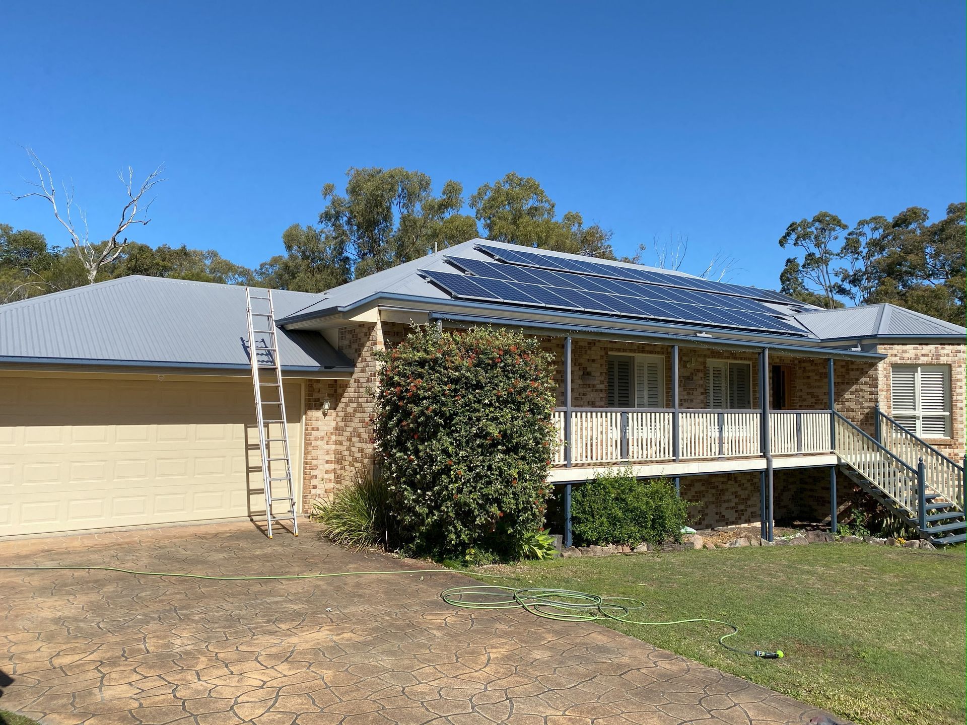 House with solar panels on the roof, a ladder, and a bush in front. Blue sky.