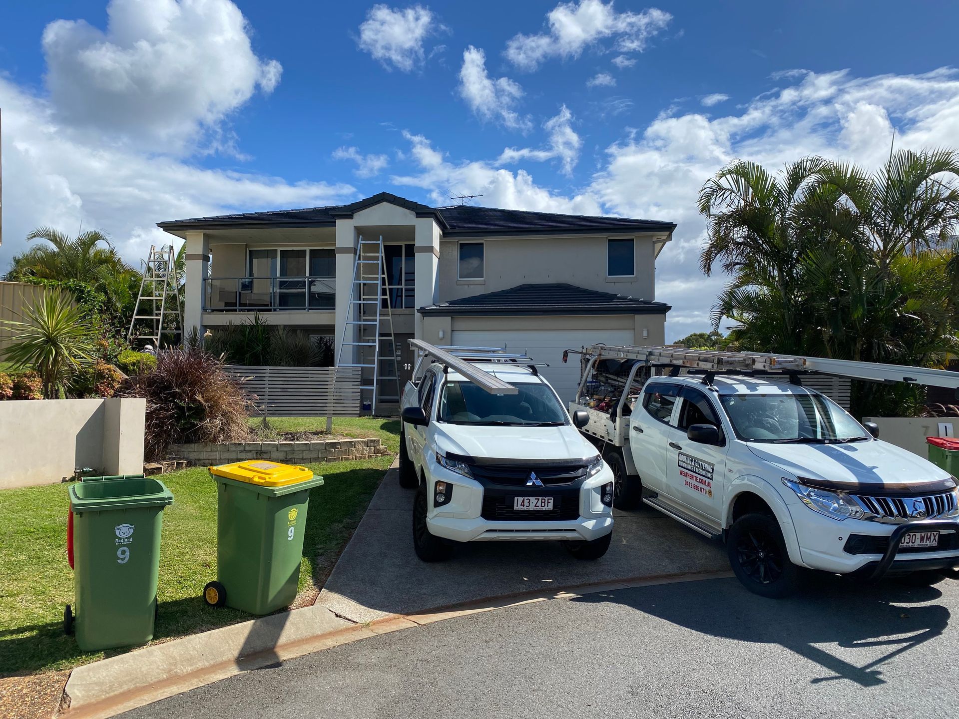 Two white trucks parked in front of a two-story house with a dark roof. Trash cans are in the foreground.