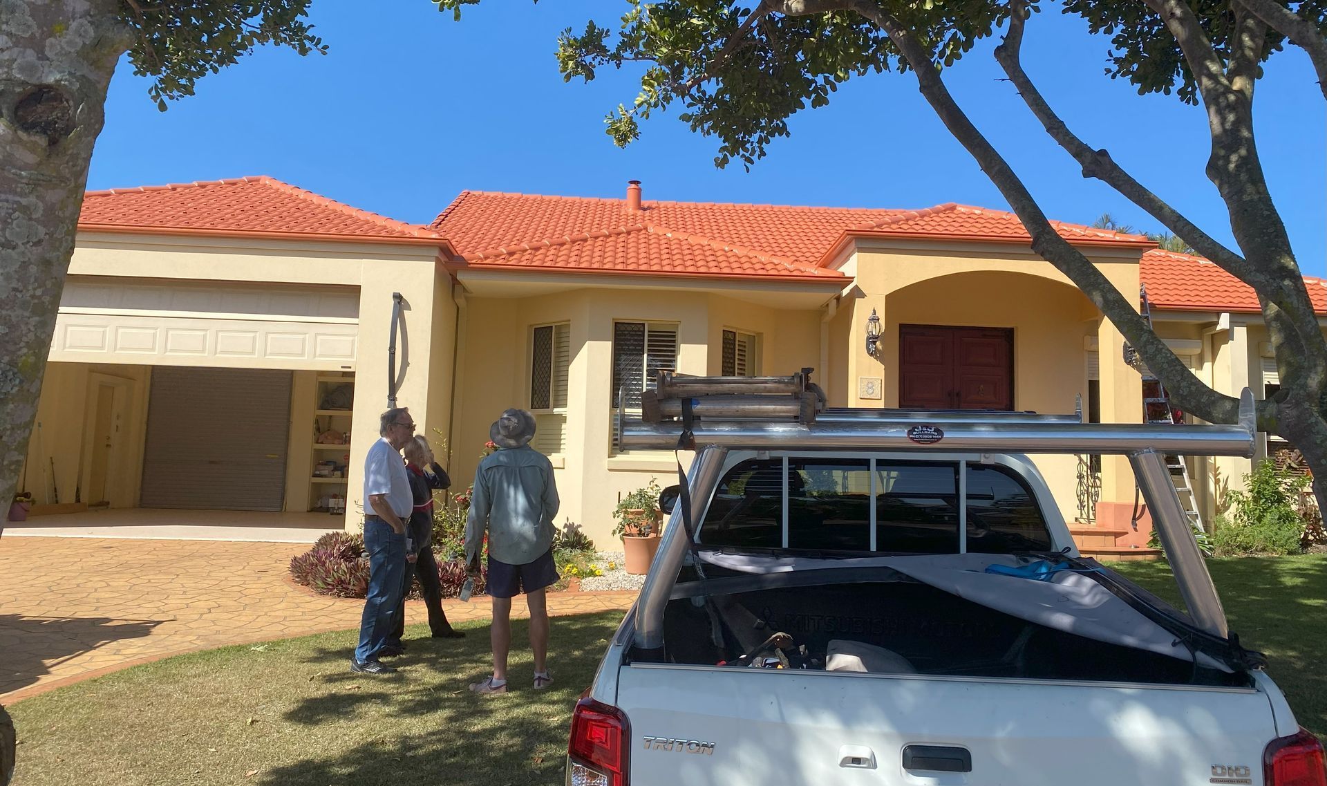Men near a beige house with orange tile roof, white pickup truck with ladder rack on the lawn.
