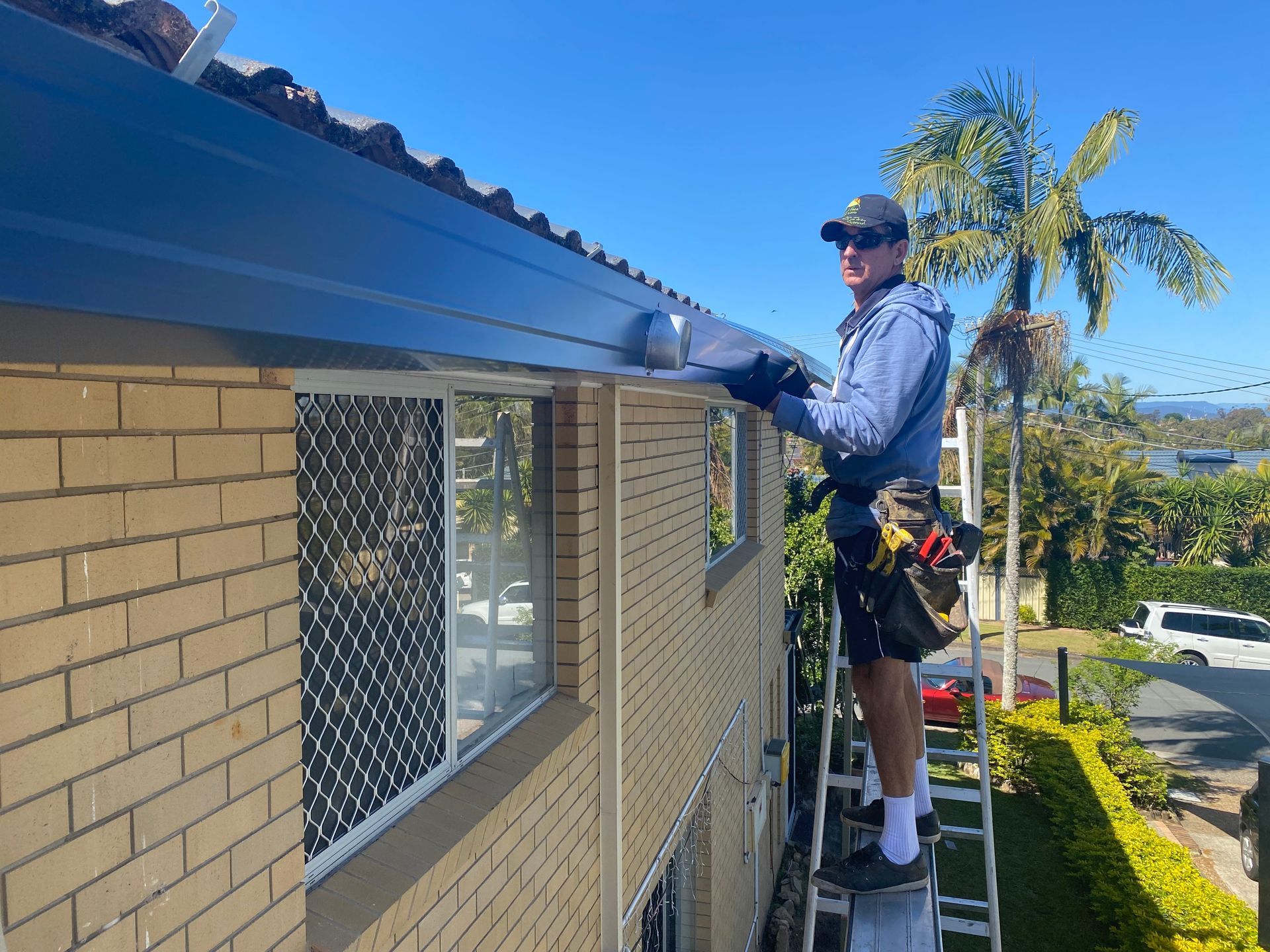 Man on ladder installing a black gutter on a brick house with a blue sky background.