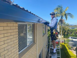 Man on ladder installing dark gutter on a brick house, sunny day.