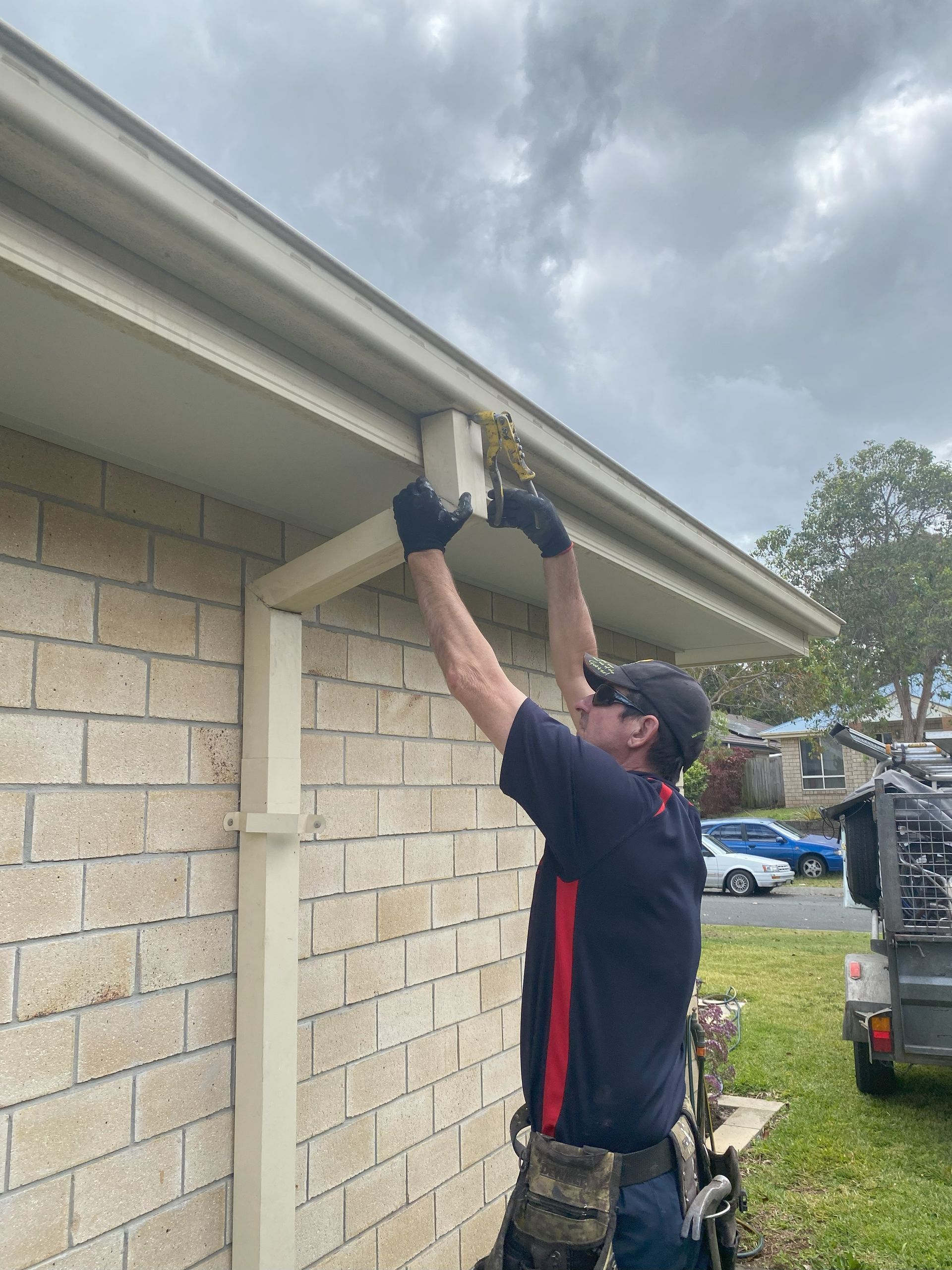 Man wearing gloves, installing fascia board under a roof eave on a brick house.