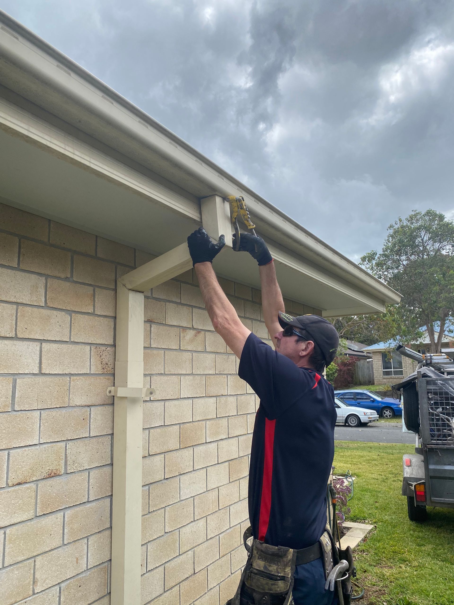 Man in gloves installing something on the eaves of a house. Brick exterior, gray sky, sunny day.