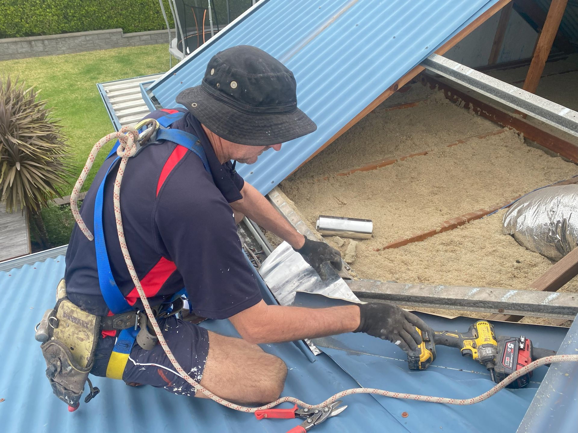 Roofer on a blue corrugated metal roof, wearing a safety harness, using a drill near insulation.