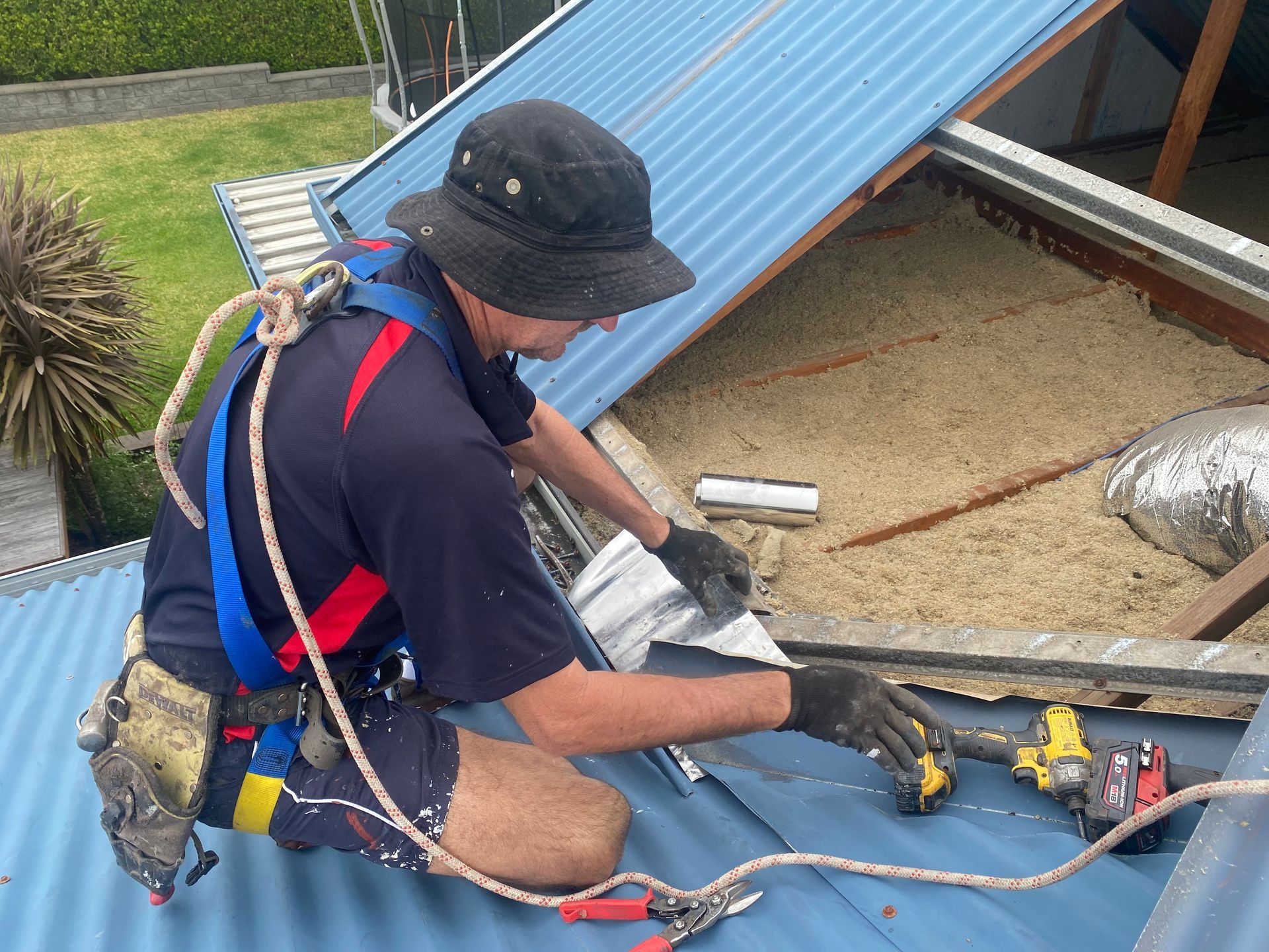 Roofer in safety harness on a blue corrugated iron roof, using a drill.