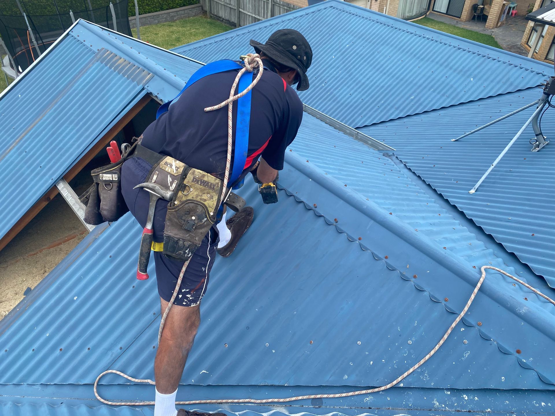 Roofer on a blue metal roof, wearing safety harness and hat, working with a drill.
