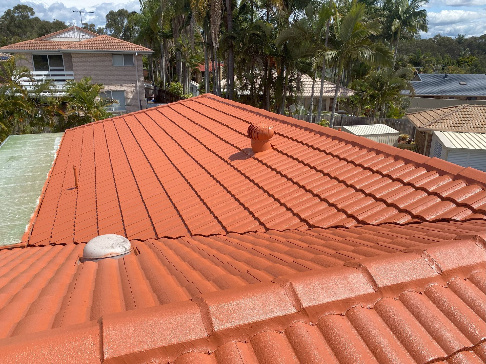 Orange tiled roof with two vents, trees and houses in the background.