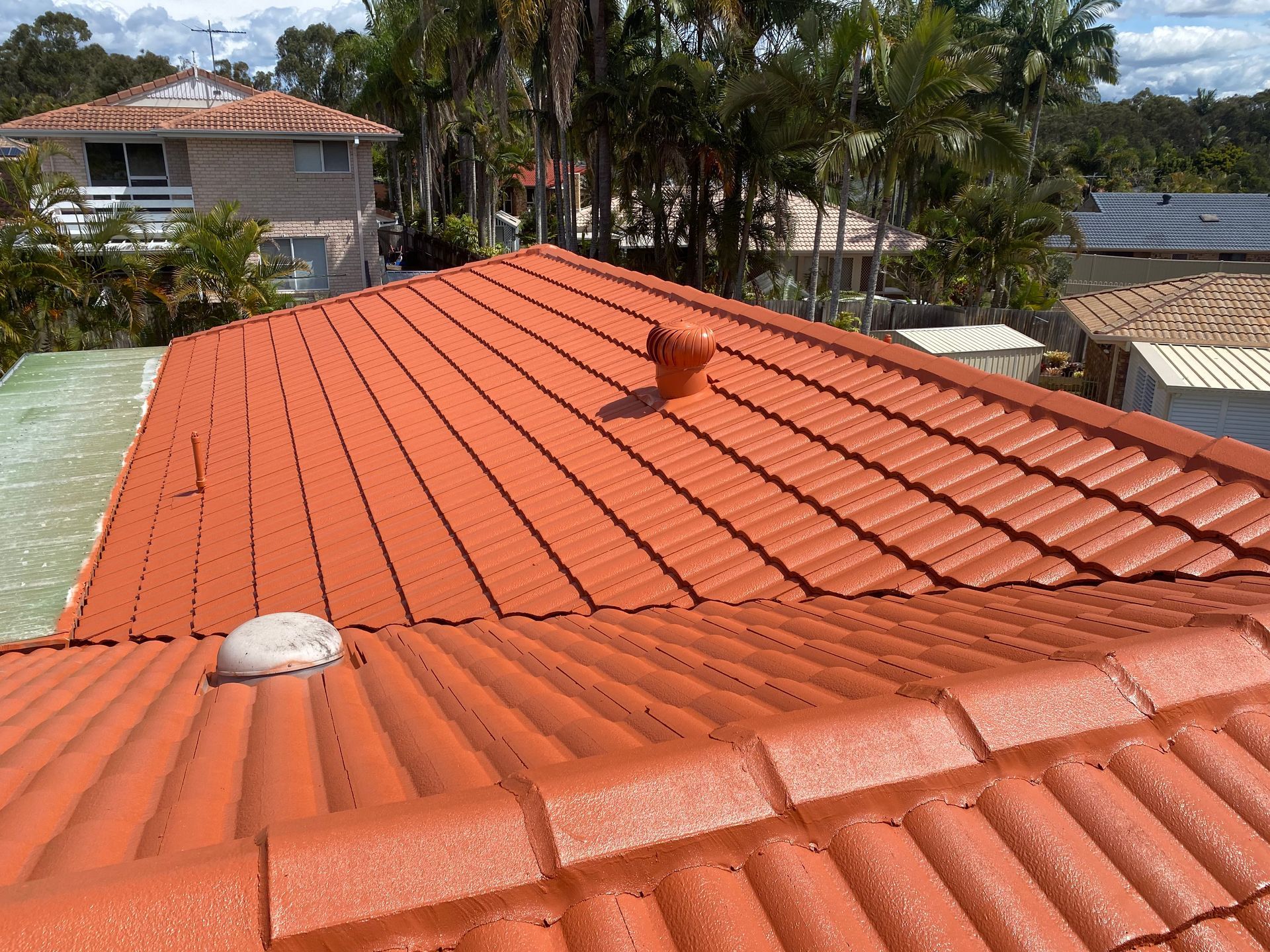 Orange tiled roof with two vents, trees, and houses in background.