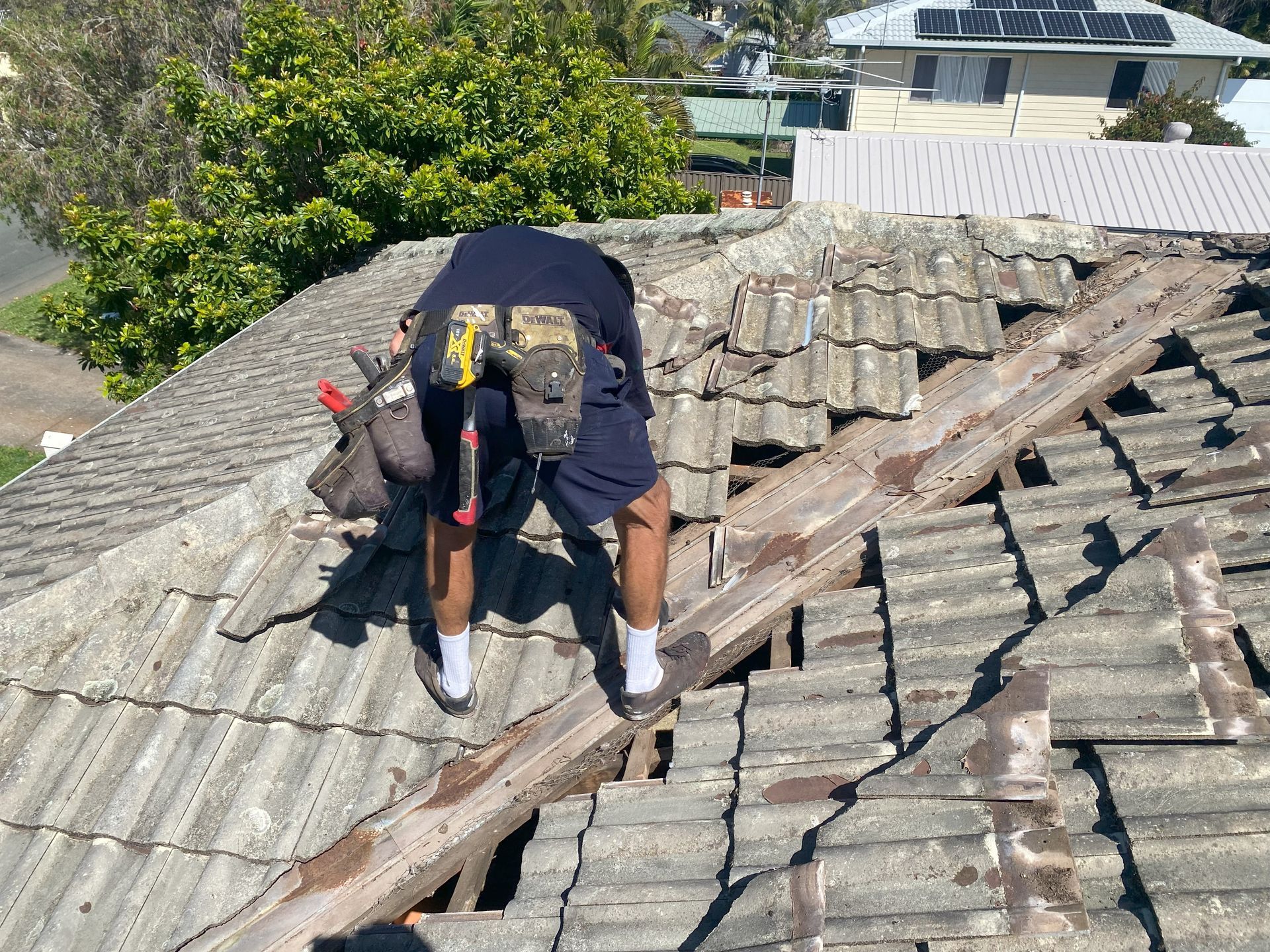 Roofer on a damaged tiled roof; he's wearing work shorts, and using tools to repair it. Sunny day.