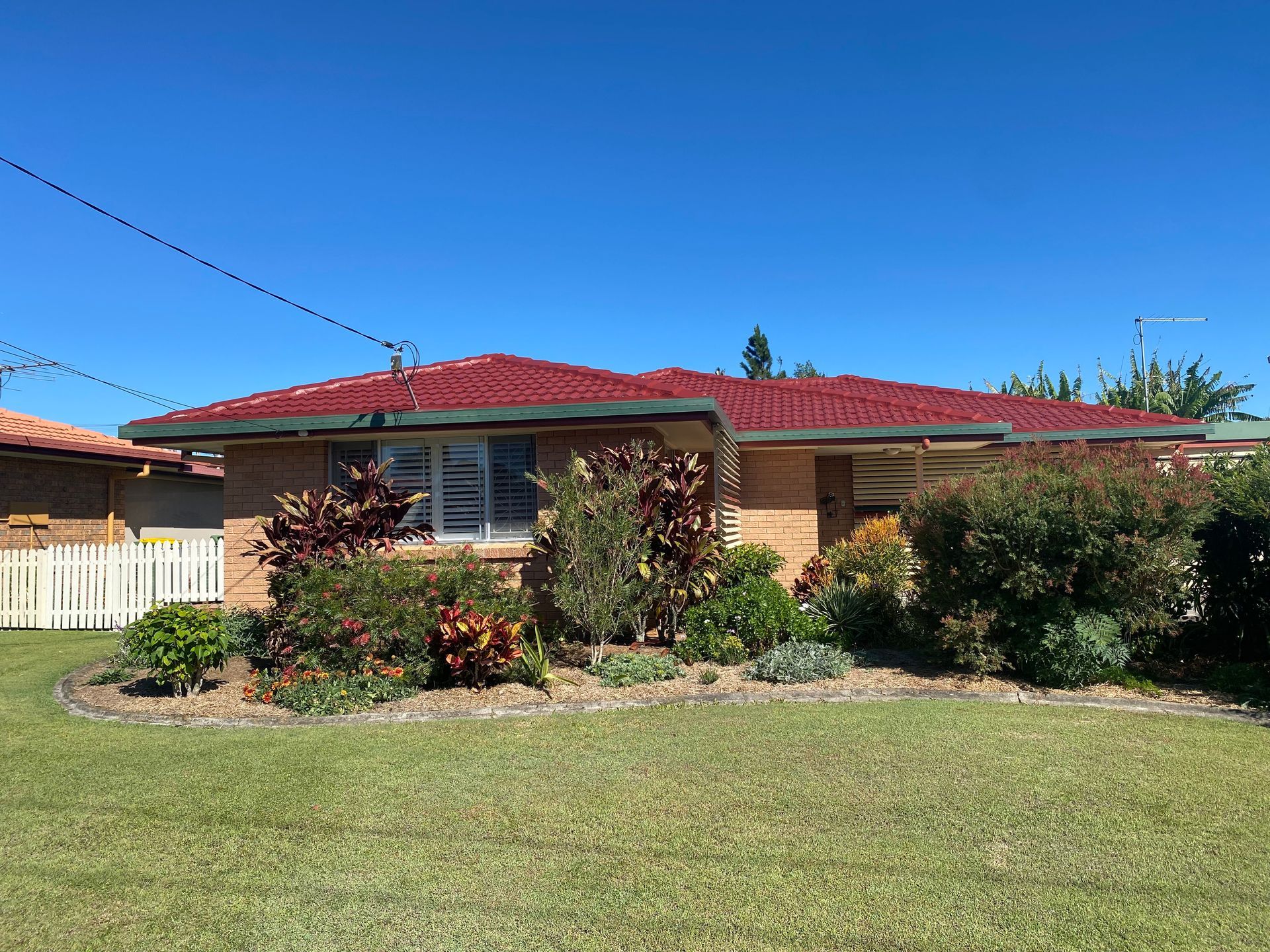 Brick home with red tiled roof, surrounded by green lawn and various bushes under a blue sky.
