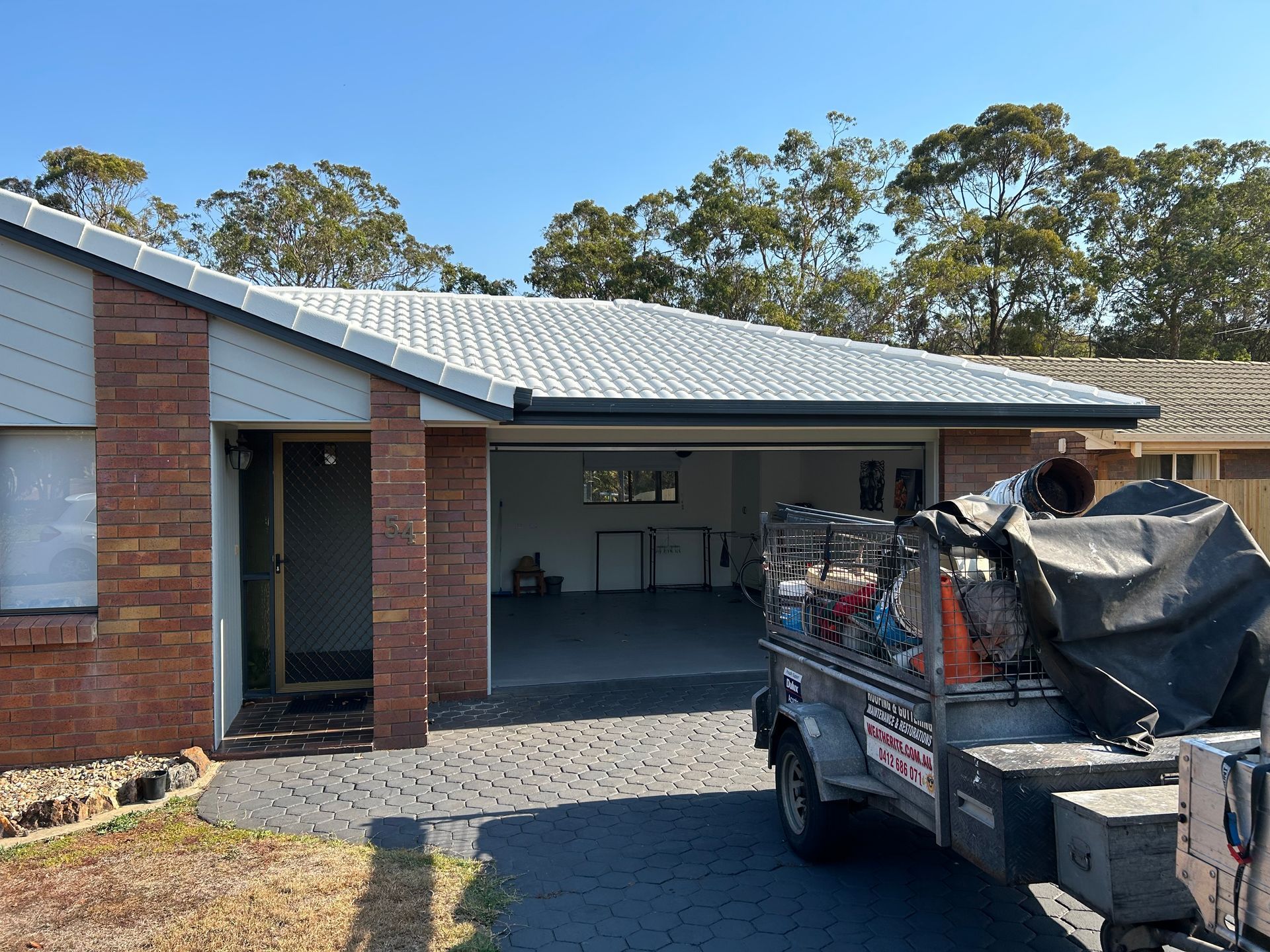 Brick house with open garage, trailer parked in front, blue sky.