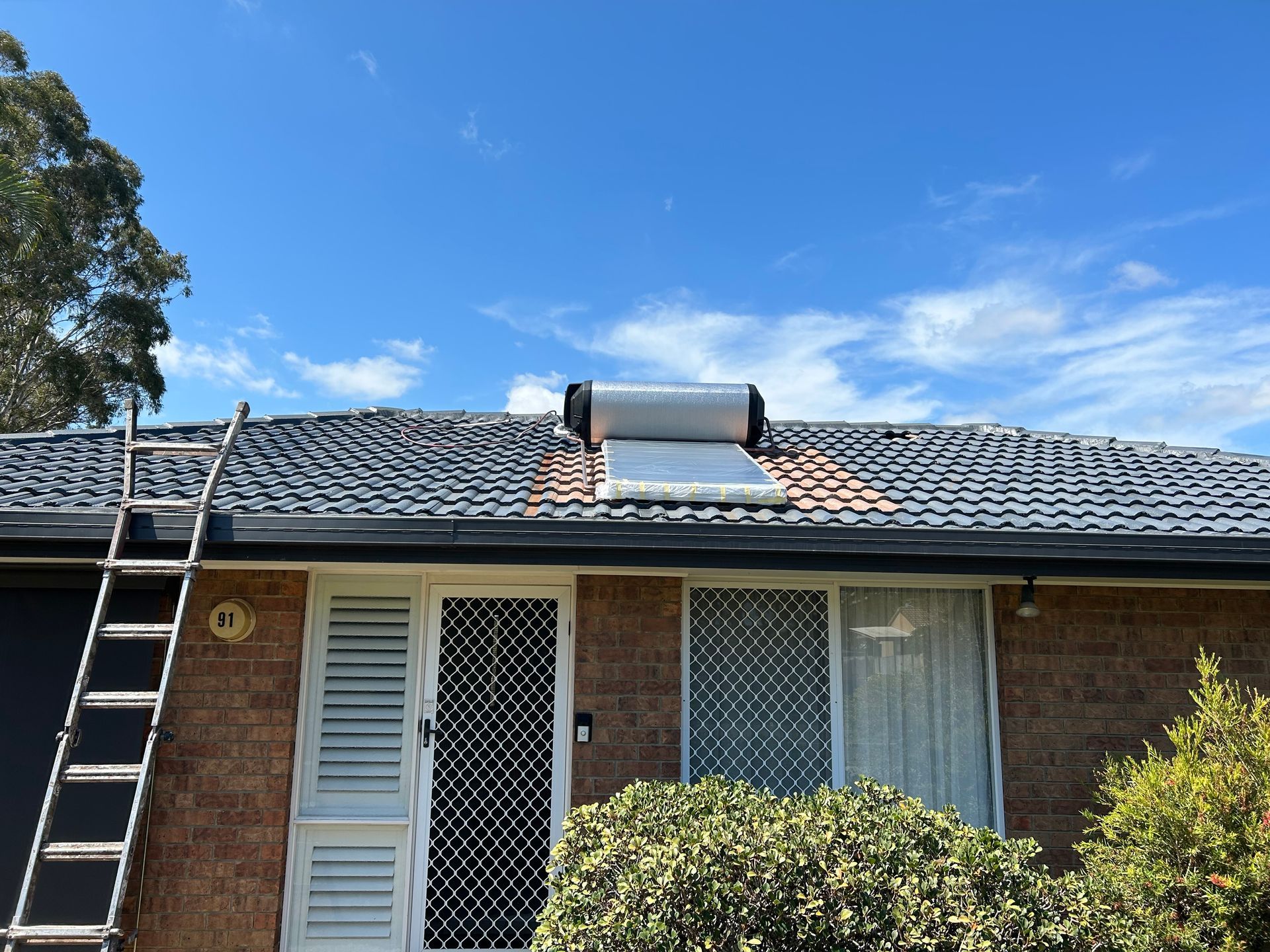 Solar water heater on roof of a brick house with a ladder leaning against the building under a blue sky.