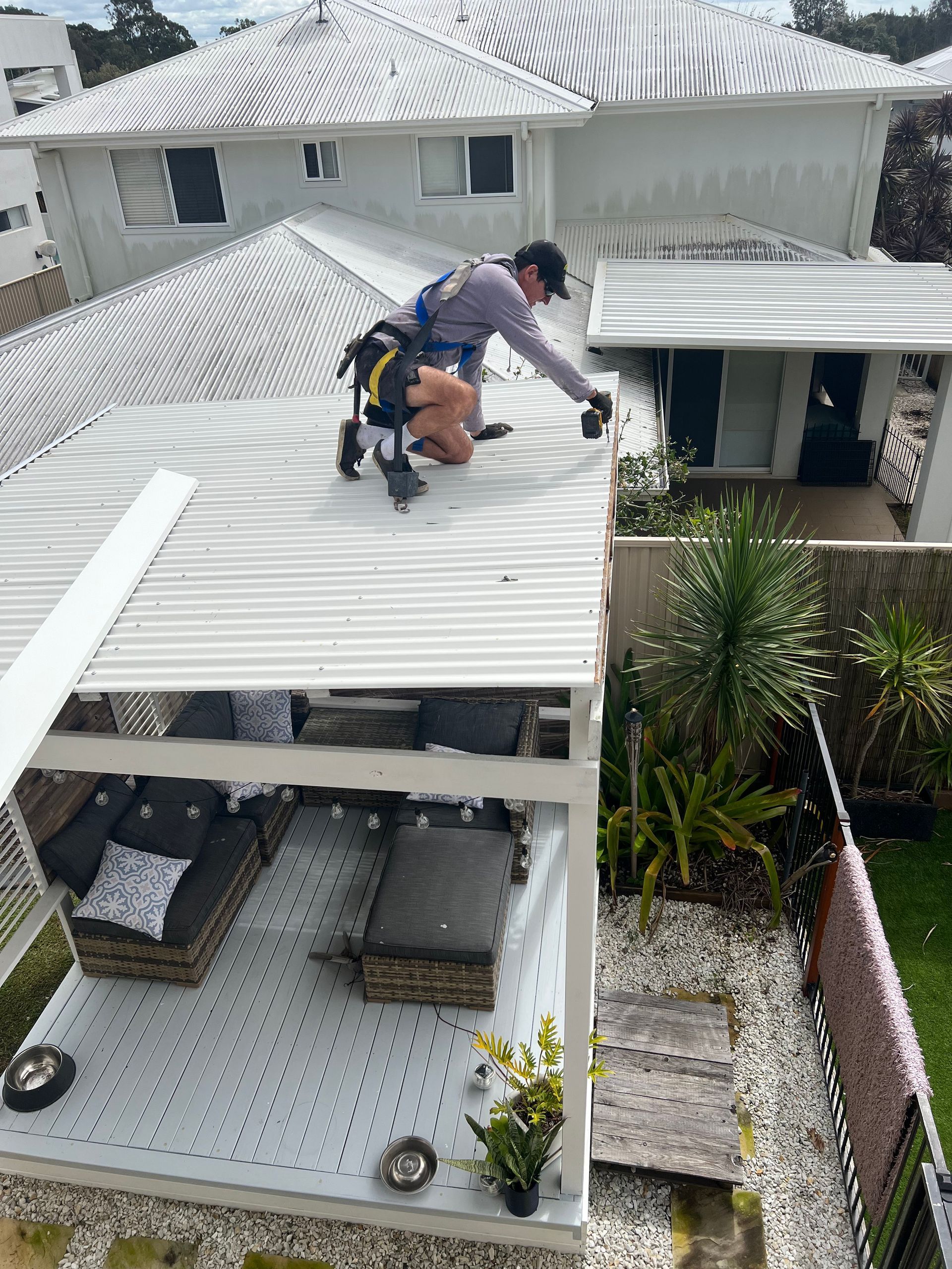 Roofer on a white corrugated roof, attached by safety harness, reaching toward the edge. Outdoor setting.