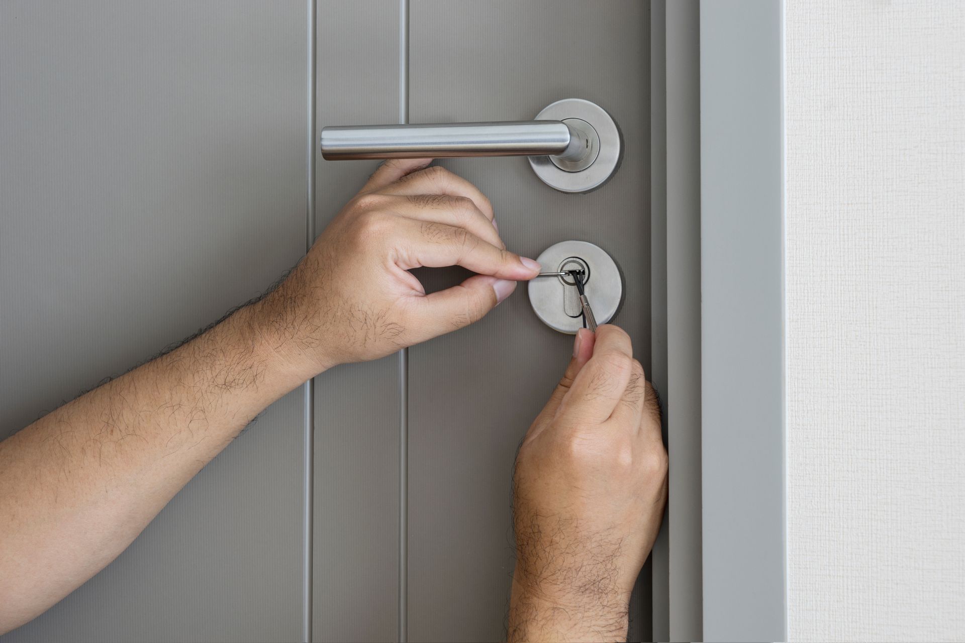 Hands using a pick and key to manipulate a door lock on a gray door.