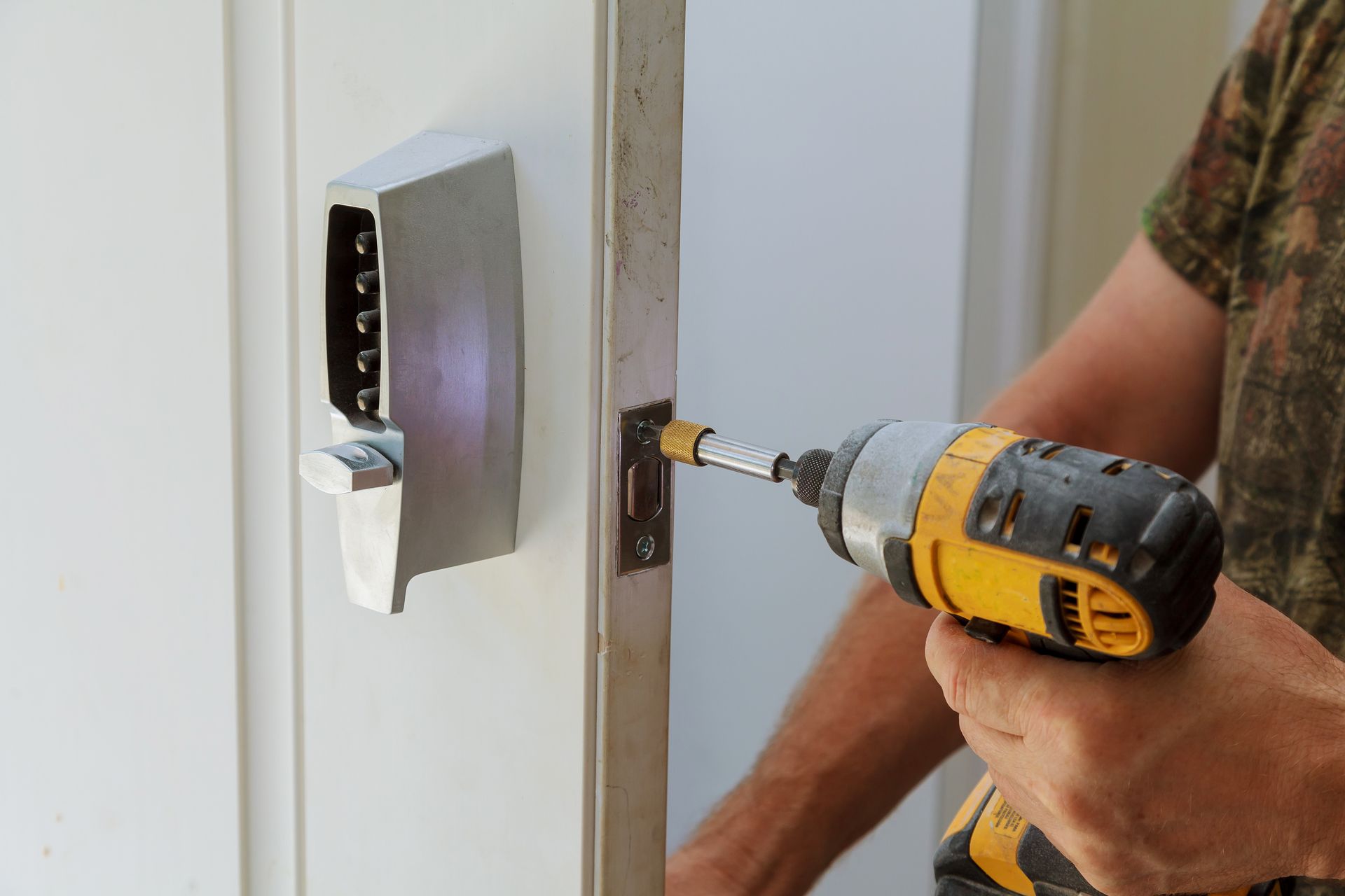 Person using a yellow drill to install a silver digital keypad door lock on a white door.